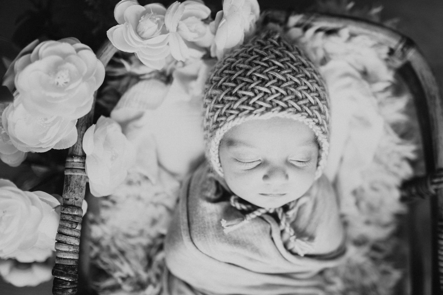 Black and white newborn photo of sleeping baby wrapped in swaddle wearing knit bonnet surrounded by white flowers.