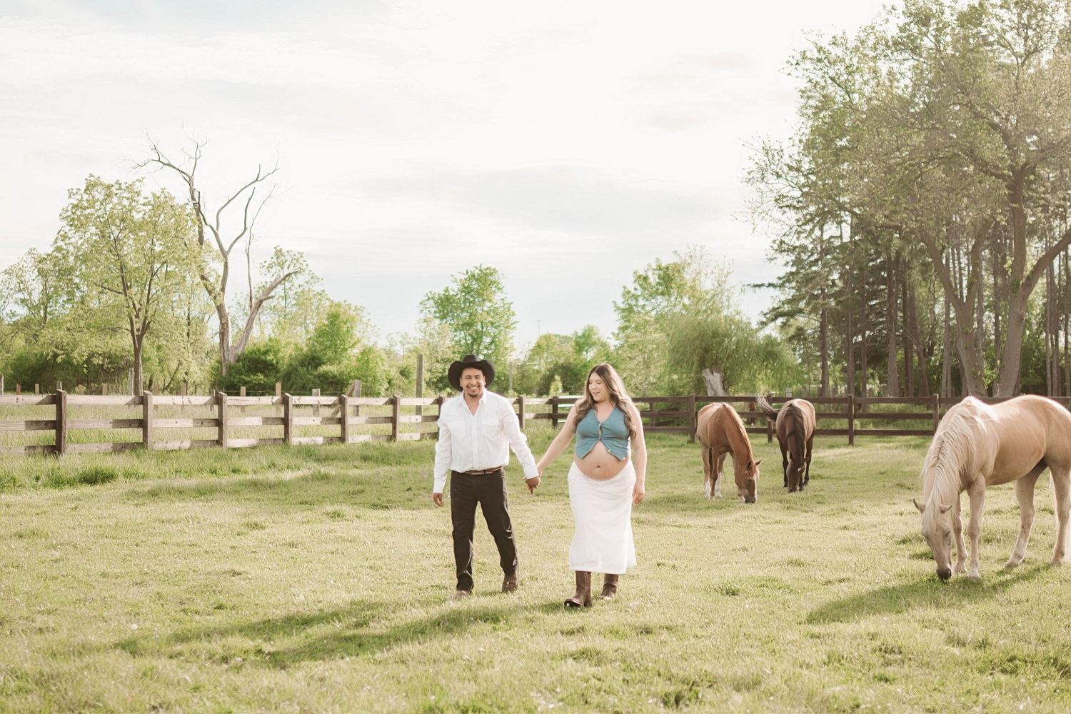 Pregnant couple holding hands walking through green pasture with horses on a sunny day at a rustic farm.