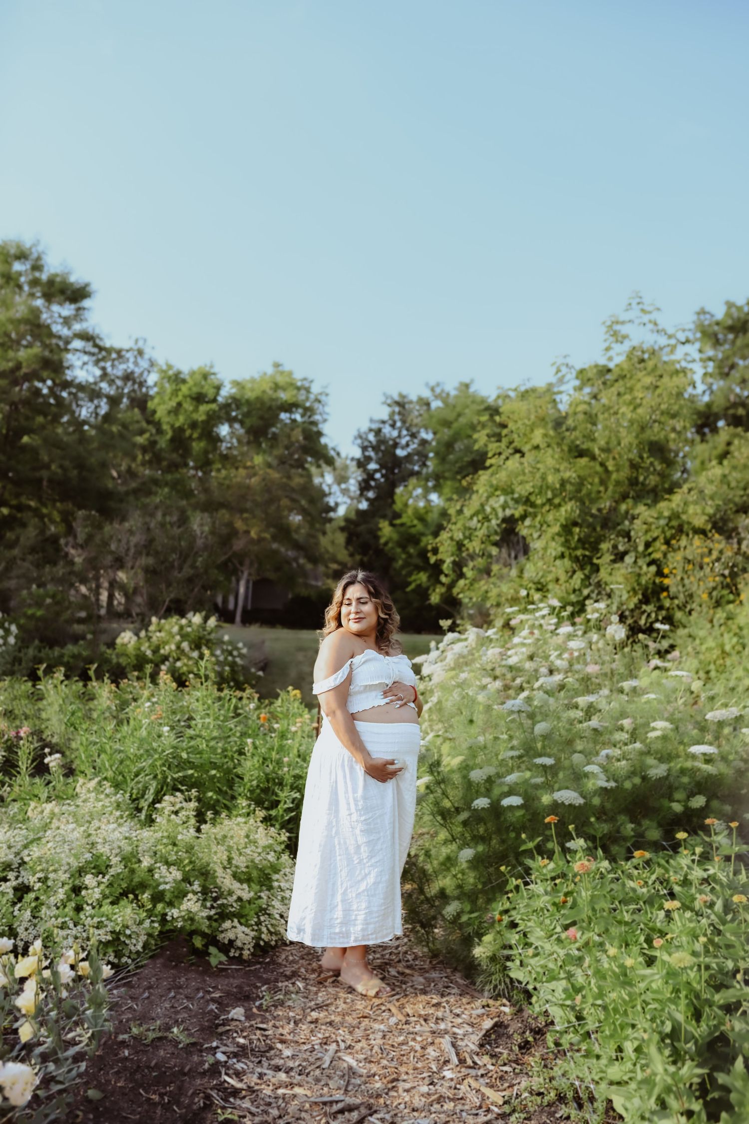 Pregnant woman in white off-shoulder dress cradling baby bump in lush garden path surrounded by wildflowers.