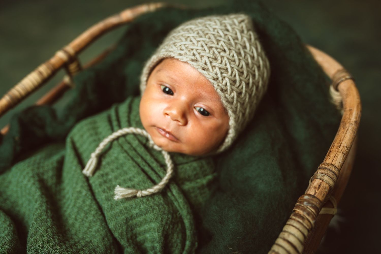 Newborn baby wrapped in green knit blanket wearing a gray bonnet hat, resting in a wicker basket.