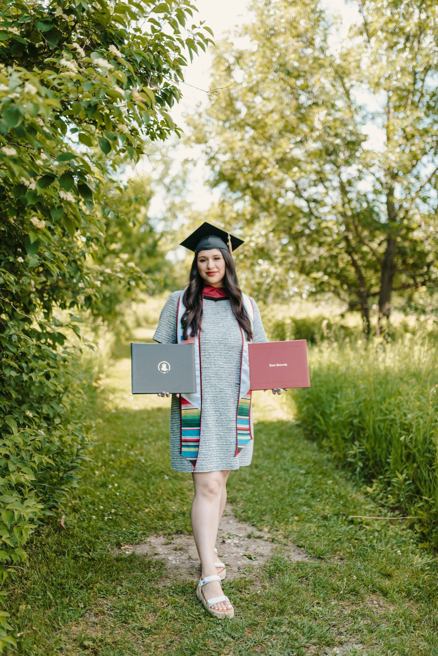 Graduate in grey dress poses with diploma on grassy outdoor path.