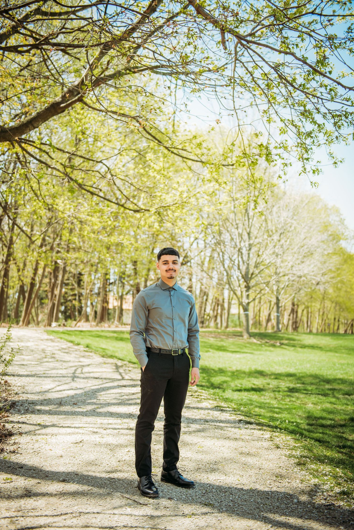 A graduate in casual attire stands on a sunlit path surrounded by spring trees.