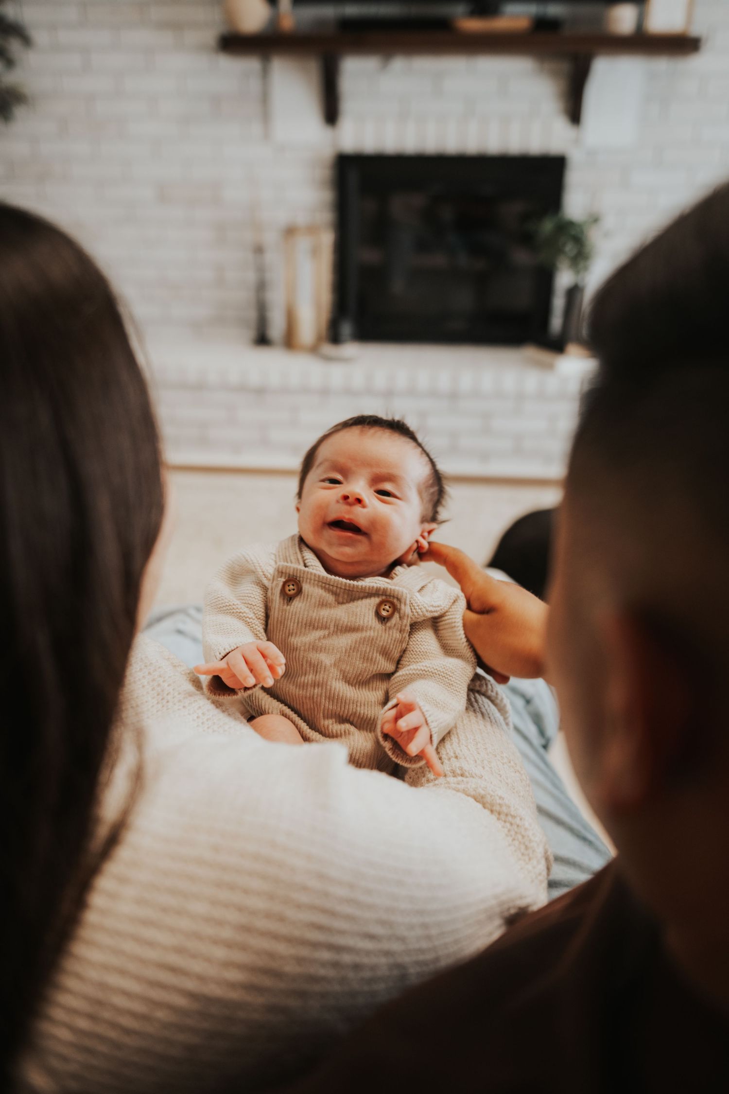 A smiling newborn baby in a beige romper being held by two parents near a white brick fireplace.