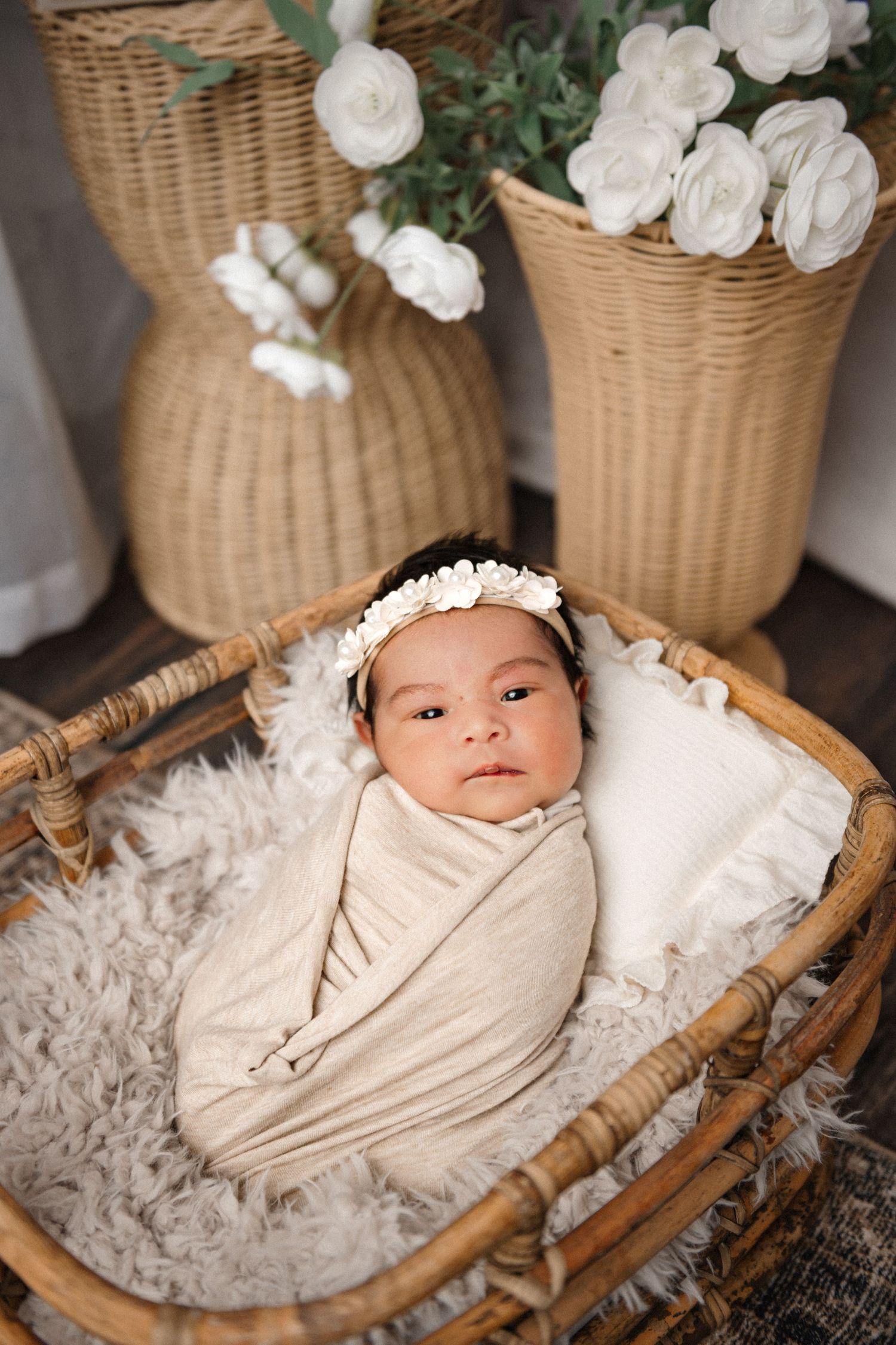 Newborn baby swaddled in cream wrap with floral headband, resting in wicker bassinet with white roses.