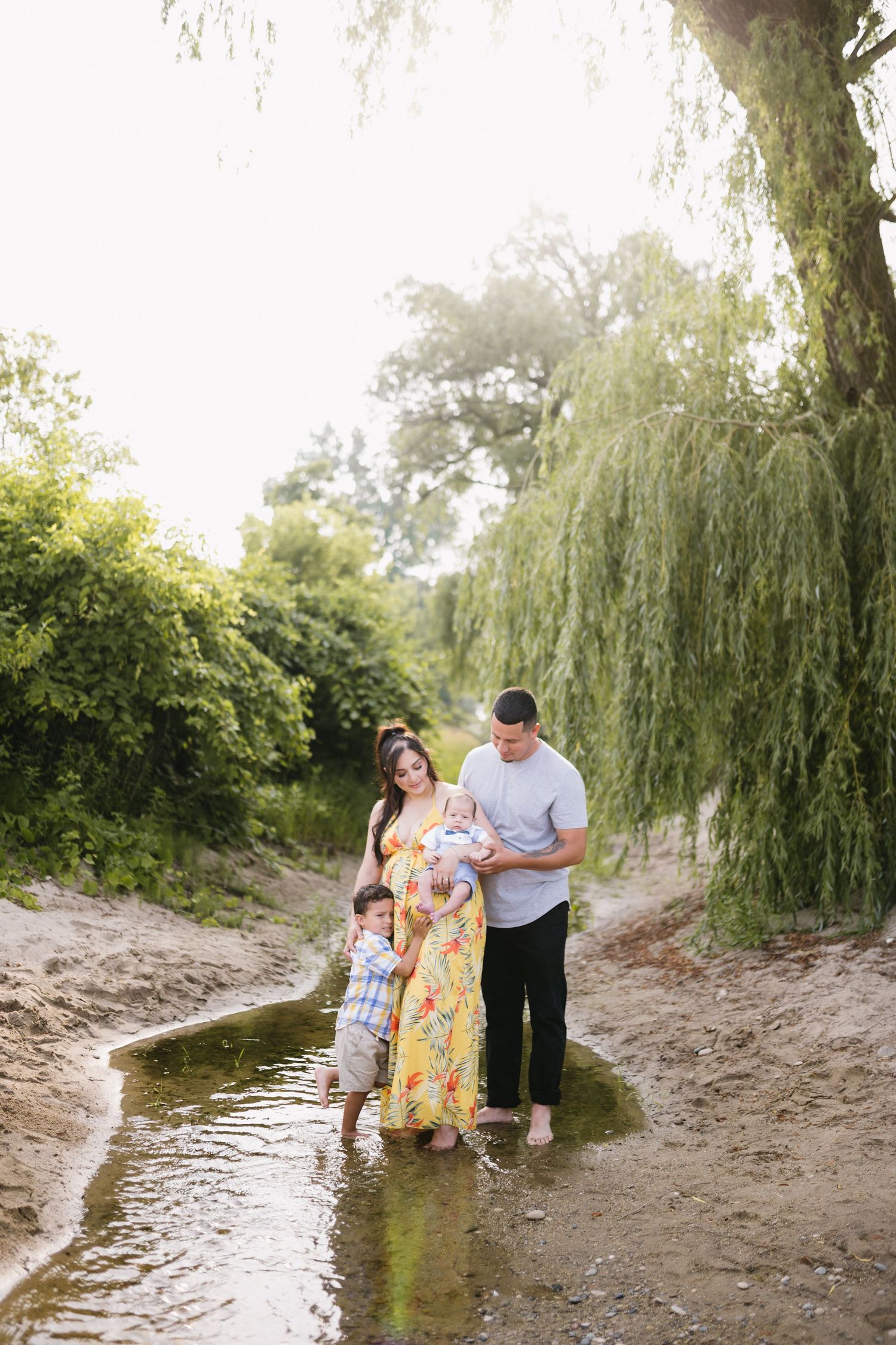 Family standing together in shallow stream beneath weeping willow trees.