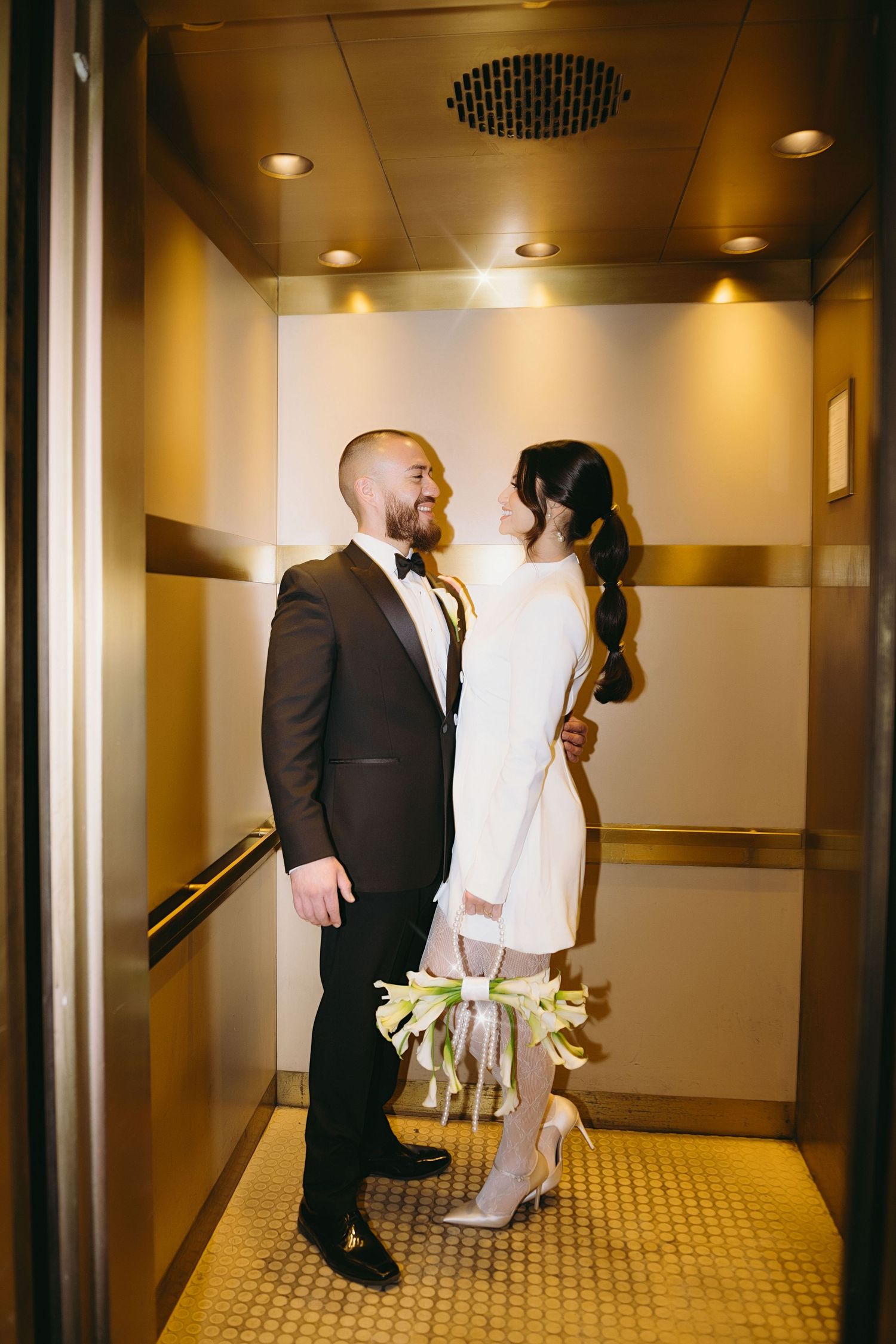 A couple walks down a warmly lit hotel hallway while carrying flowers and dressed in formal attire.