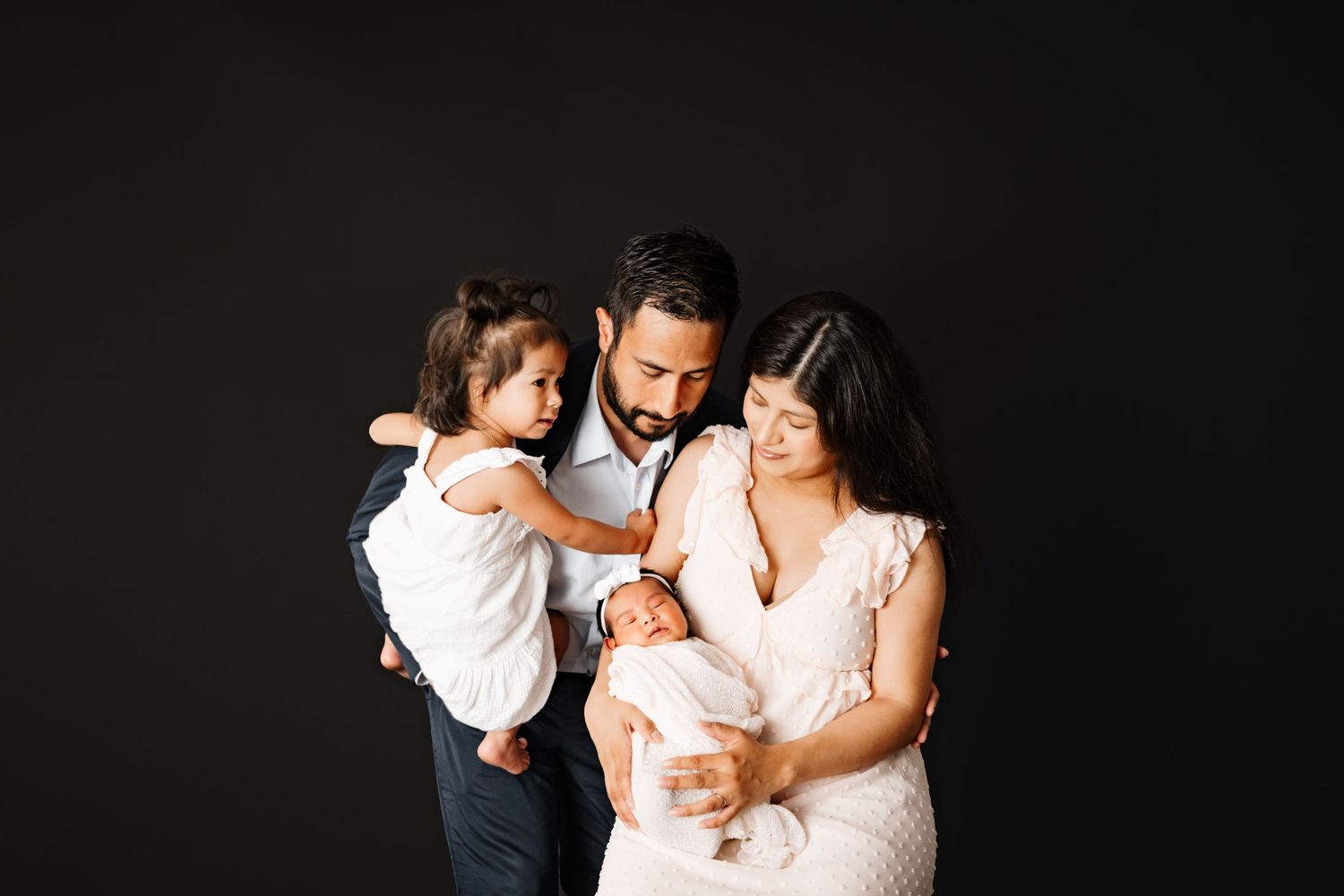 Family of four posing for a studio portrait, parents lovingly gazing at newborn baby while toddler is held close.