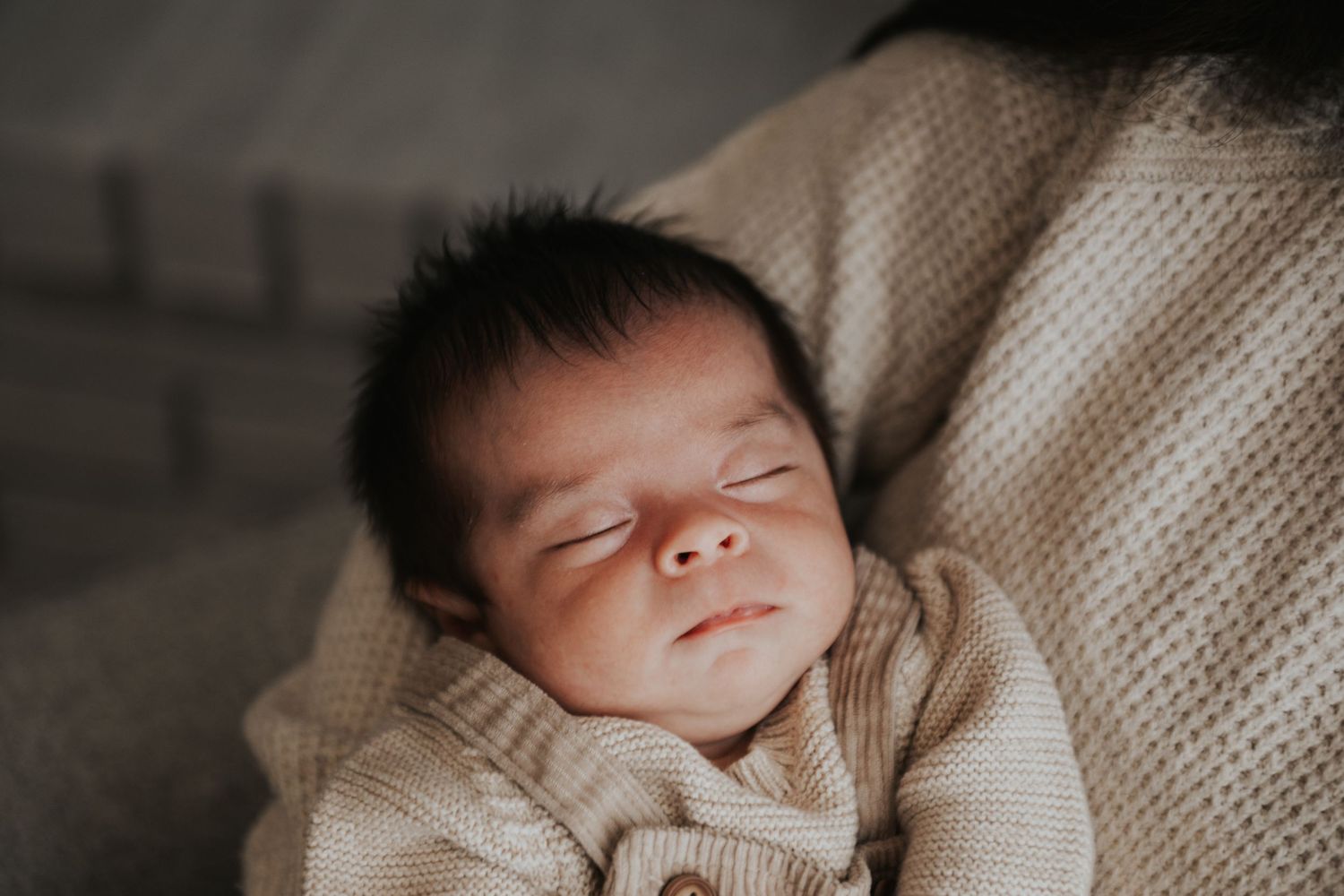 Peaceful newborn baby sleeping wrapped in a soft beige knit blanket with dark hair and delicate features.