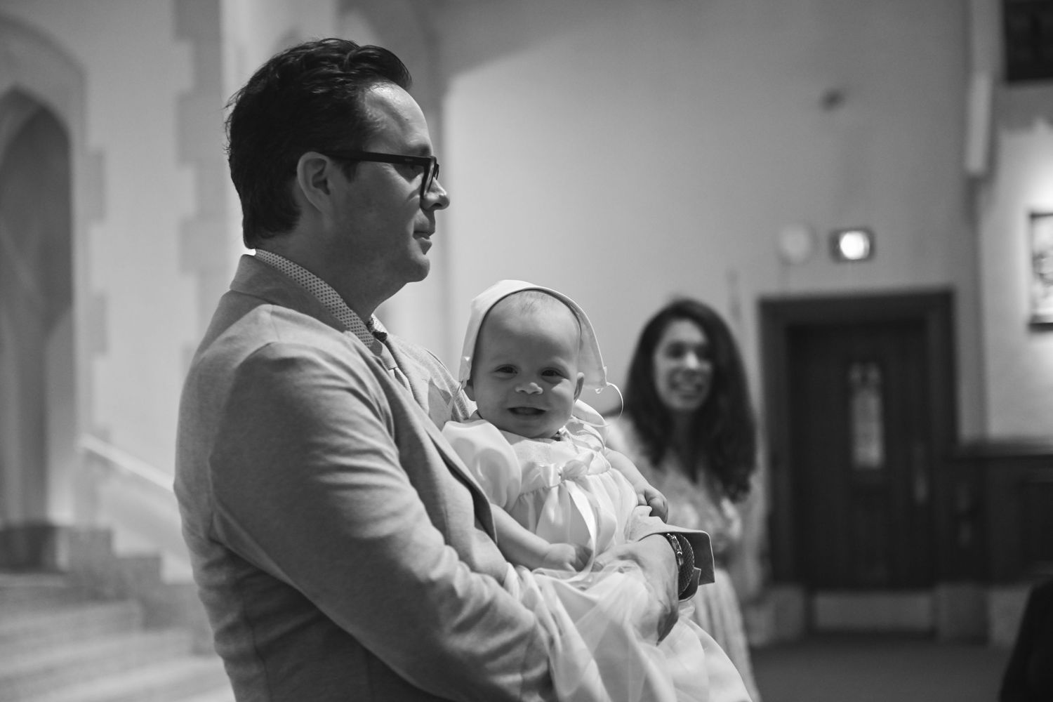 A black and white photo showing a baby being held in a church hallway.