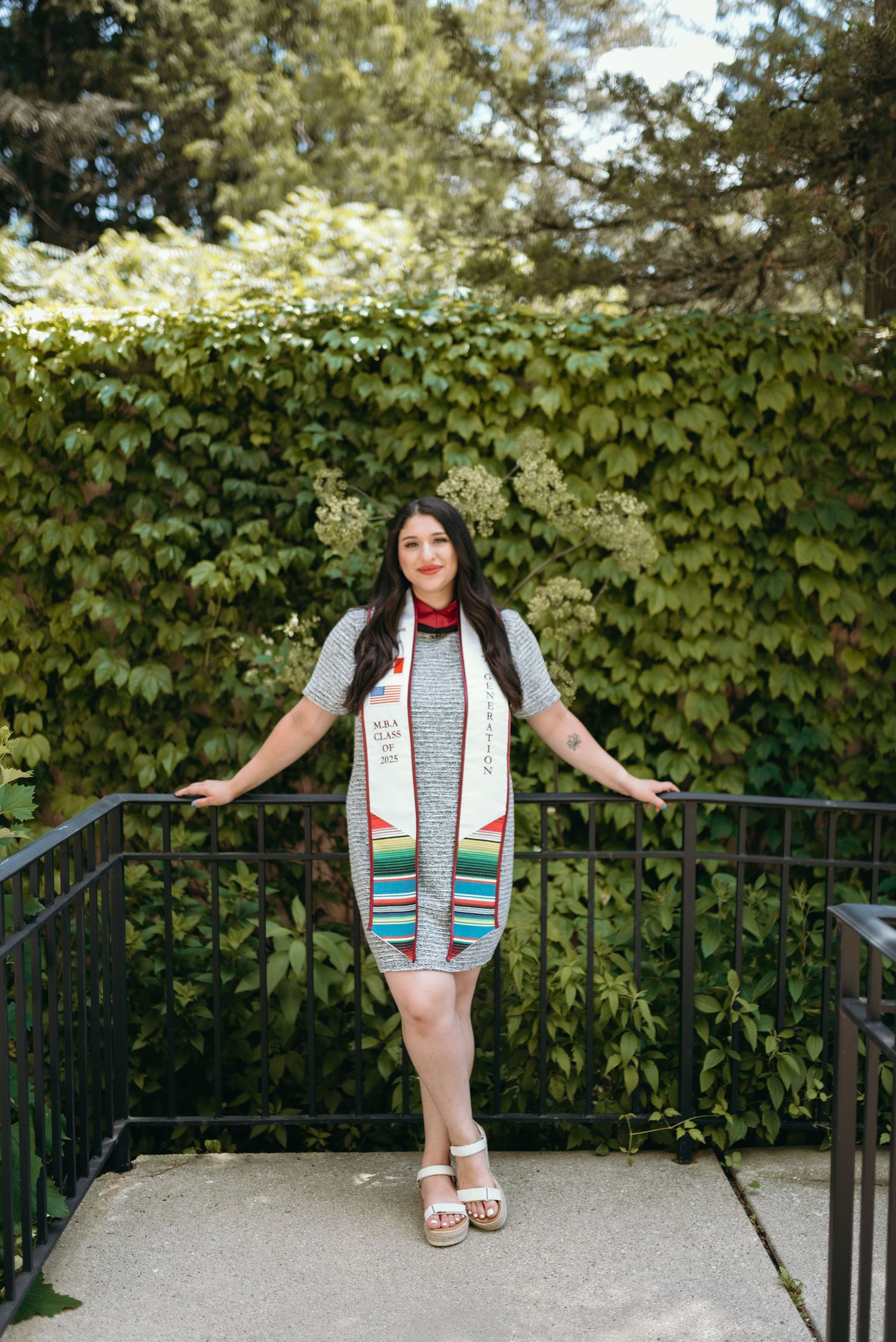 Graduate in grey dress poses playfully on metal railing during outdoor photo session.