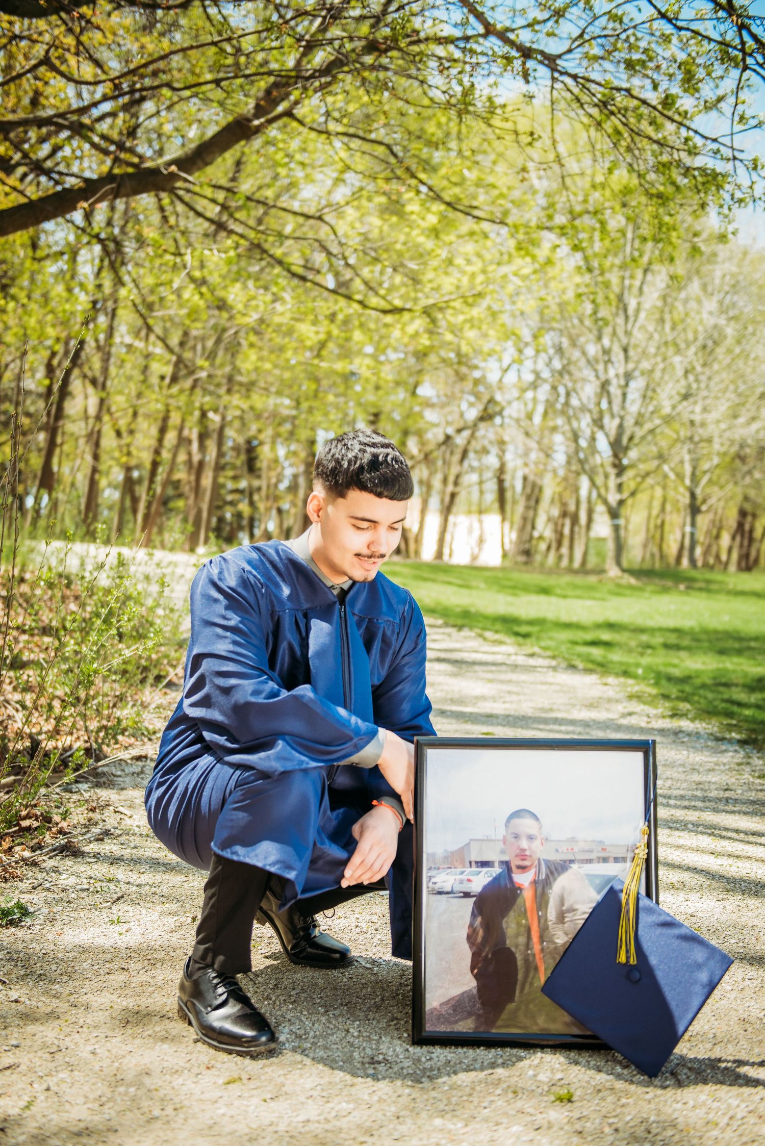 A graduate in blue regalia kneels beside graduation photos displayed on a path.
