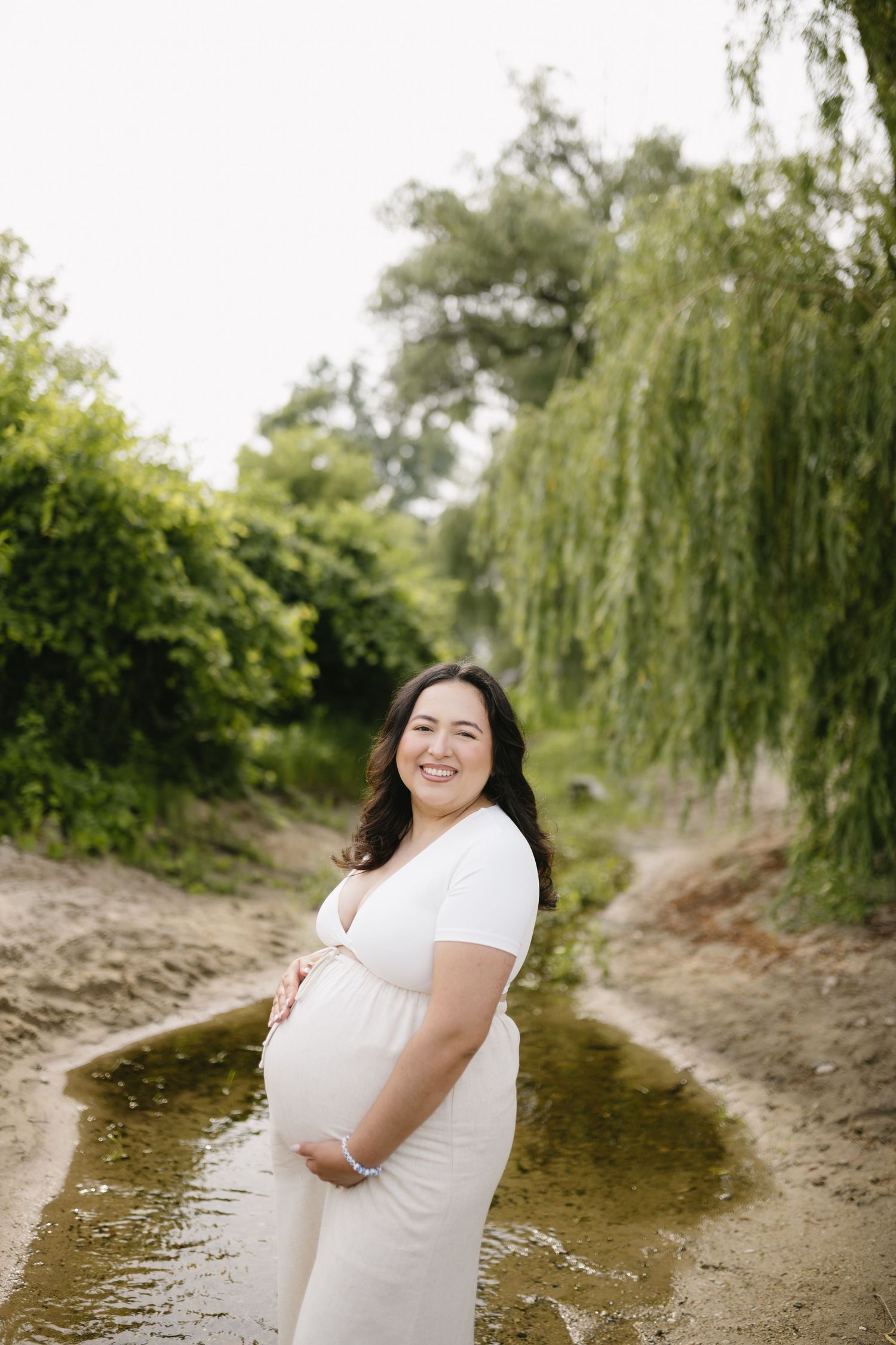 Expectant mother in white dress smiling while standing on a nature path with willow trees.