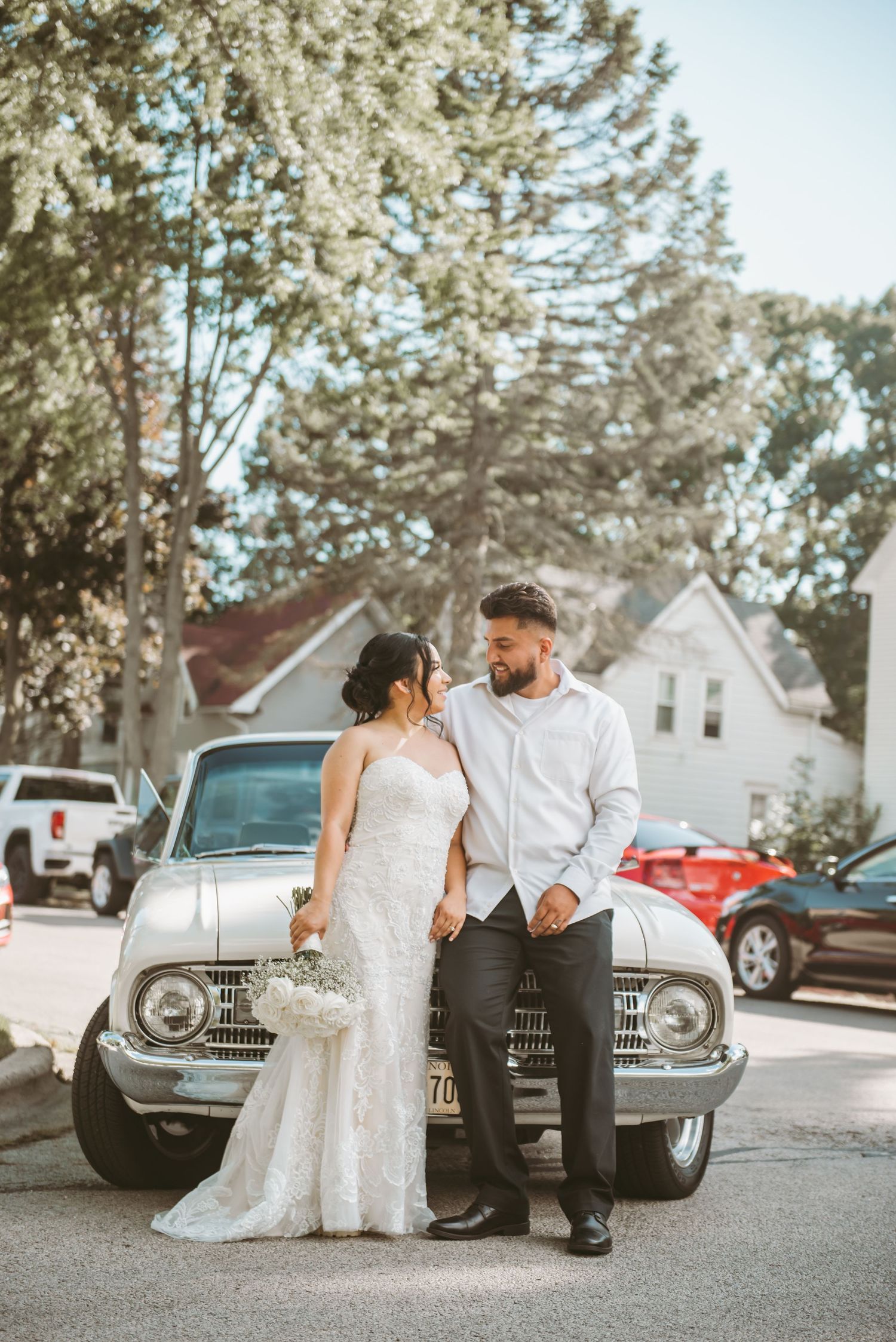 A couple shares intimate moments while sitting beside a vintage car in a suburban setting.