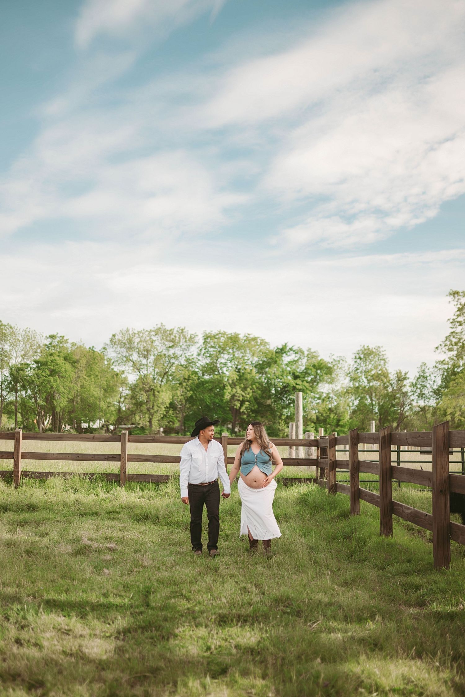 Pregnant couple walking along a wooden fence in a green meadow under a partly cloudy sky.