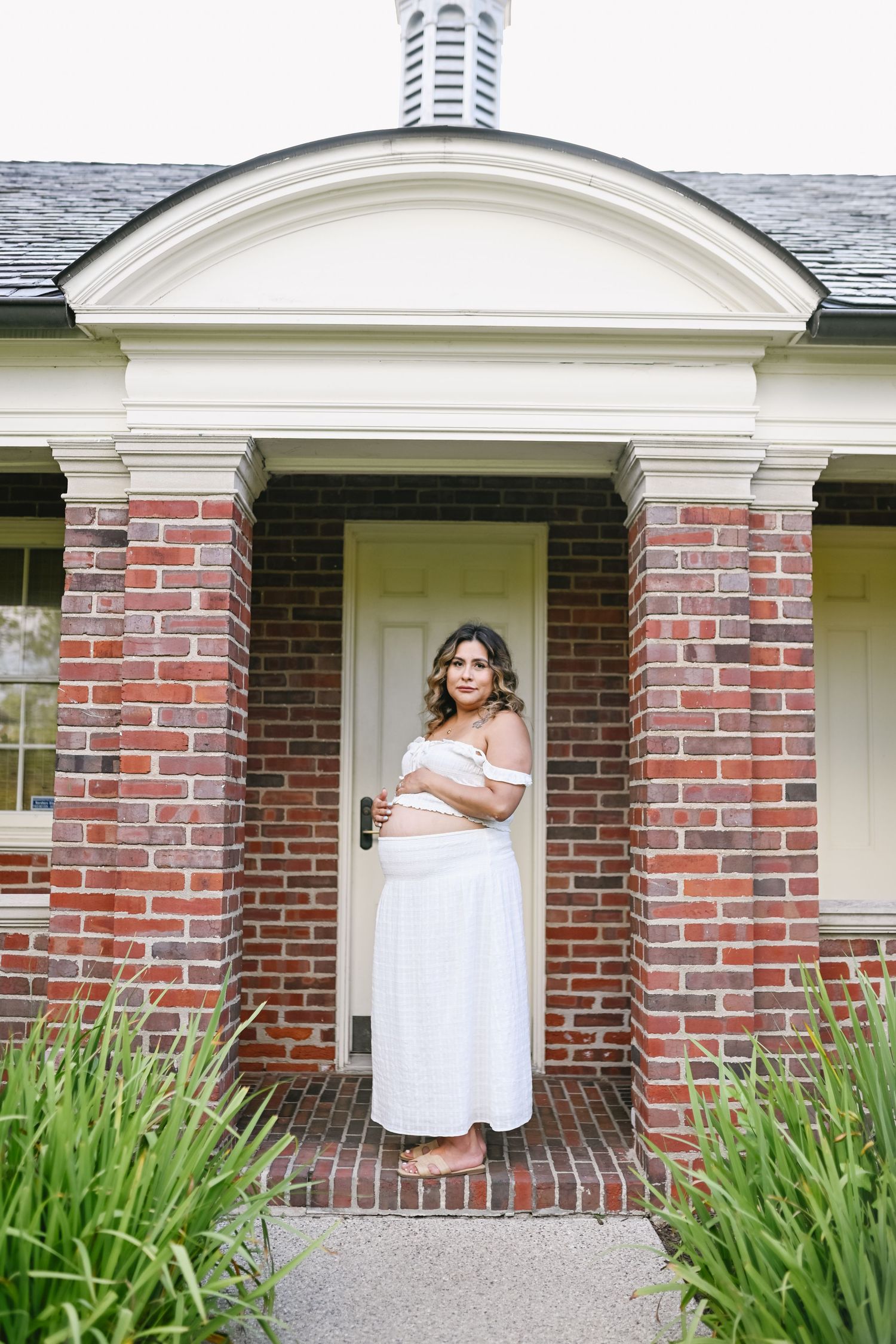 Pregnant woman in white off-shoulder dress posing at brick building entrance with white door and green plants.