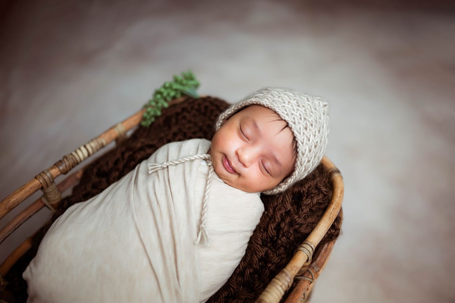 Sleeping newborn baby wrapped in cream swaddle wearing knit bonnet resting in a wicker basket with brown fur lining.