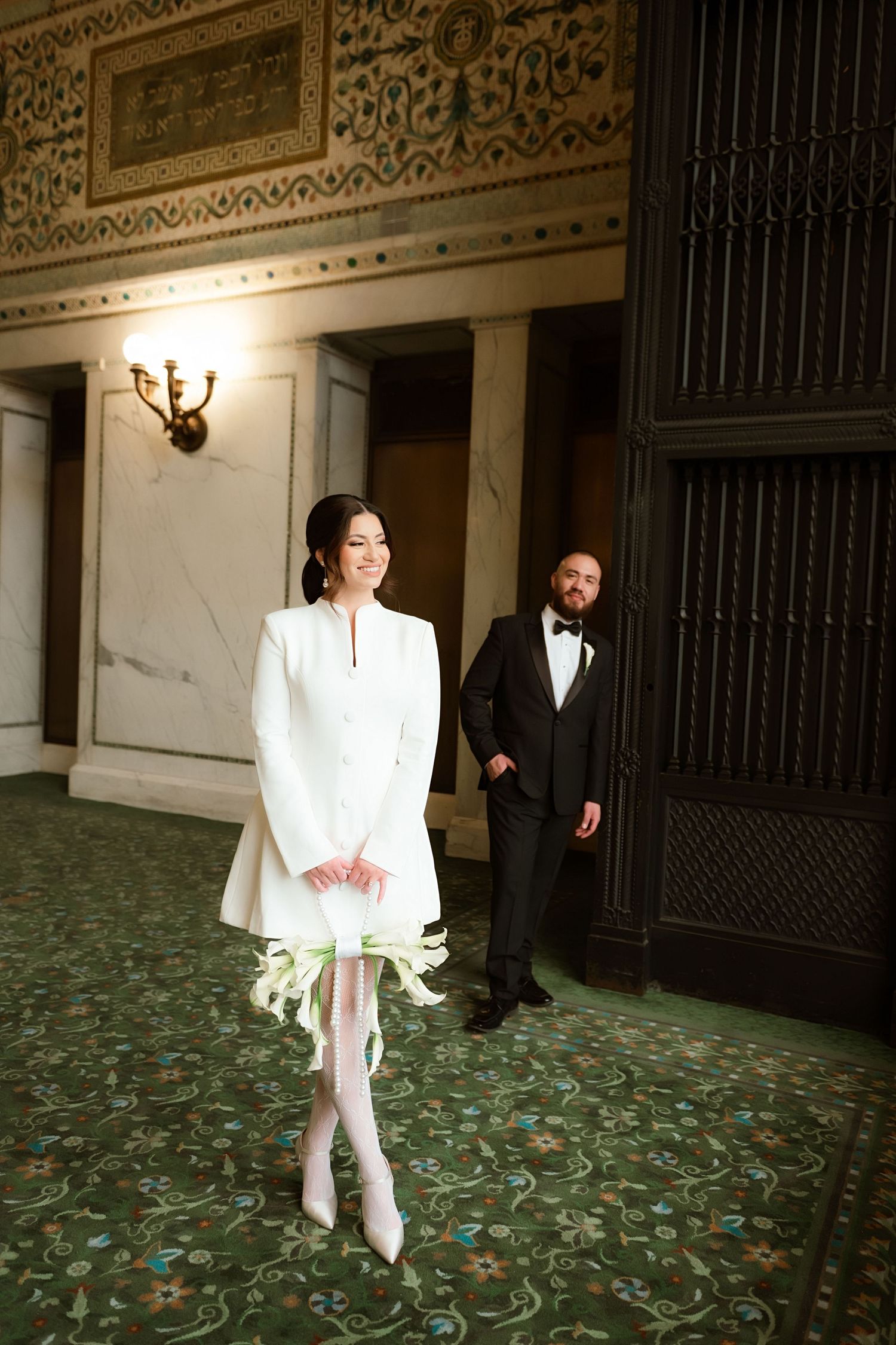 A couple in formal wedding attire poses in an elegant vintage hallway with ornate decor and patterned carpet.