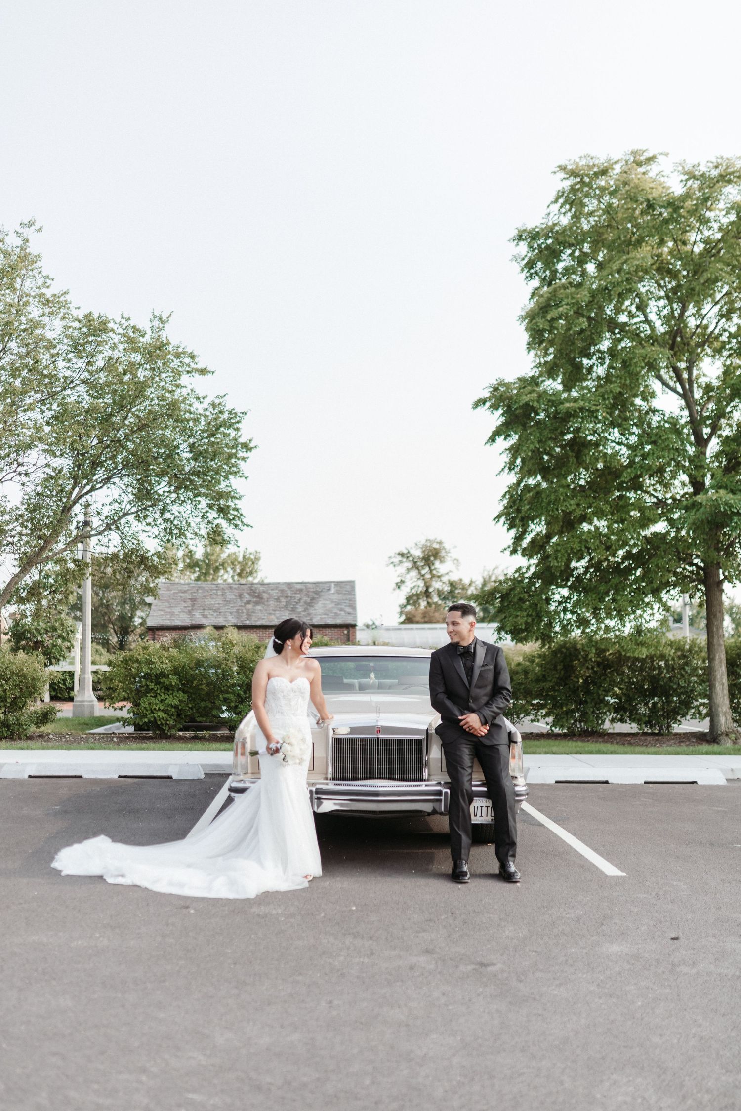 Wedding couple poses in a parking lot with trees and buildings in the background on their special day.
