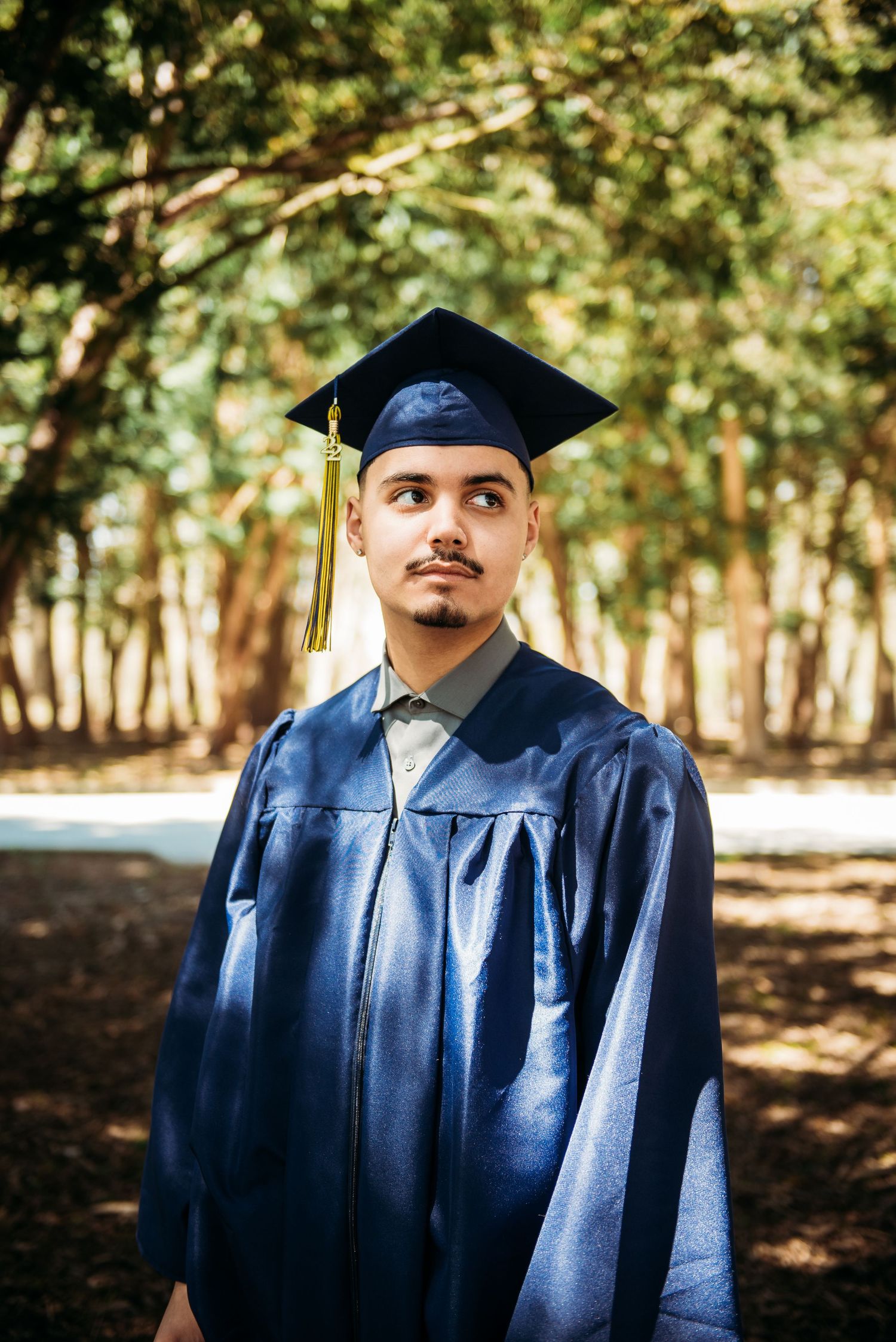 A graduate poses in traditional blue academic regalia outdoors among trees.