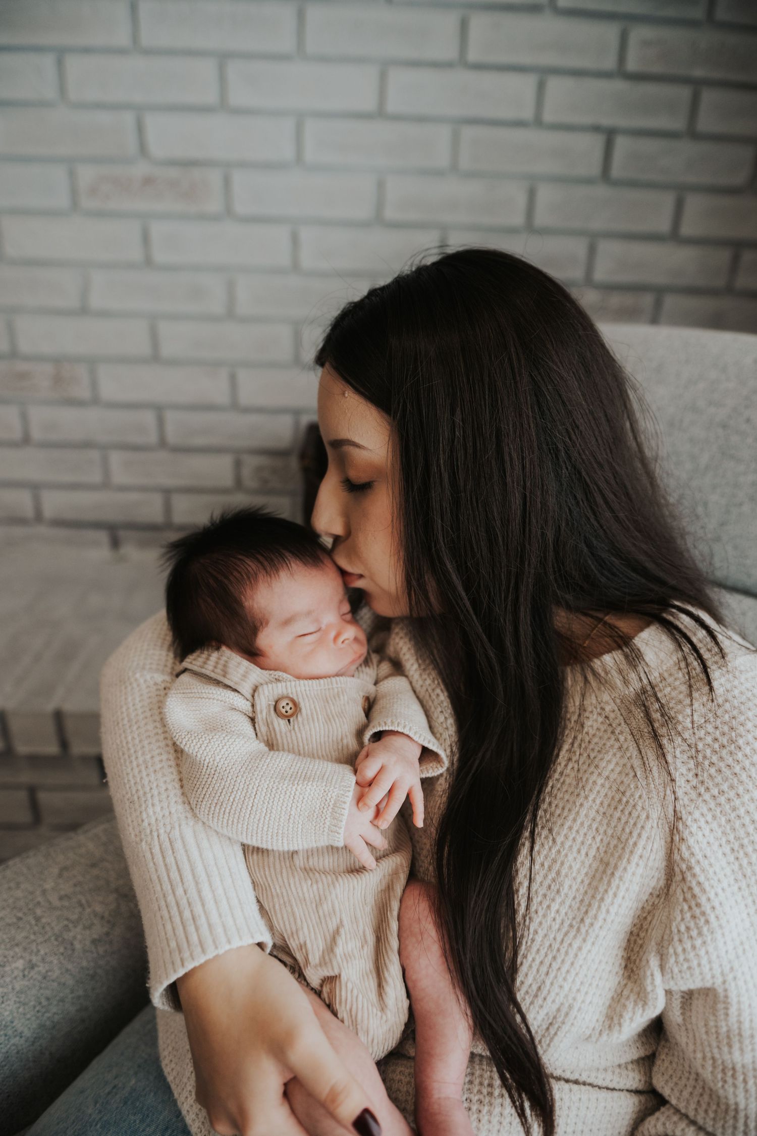 A dark-haired mother gently kisses her sleeping newborn baby dressed in a beige knit outfit while sitting indoors.