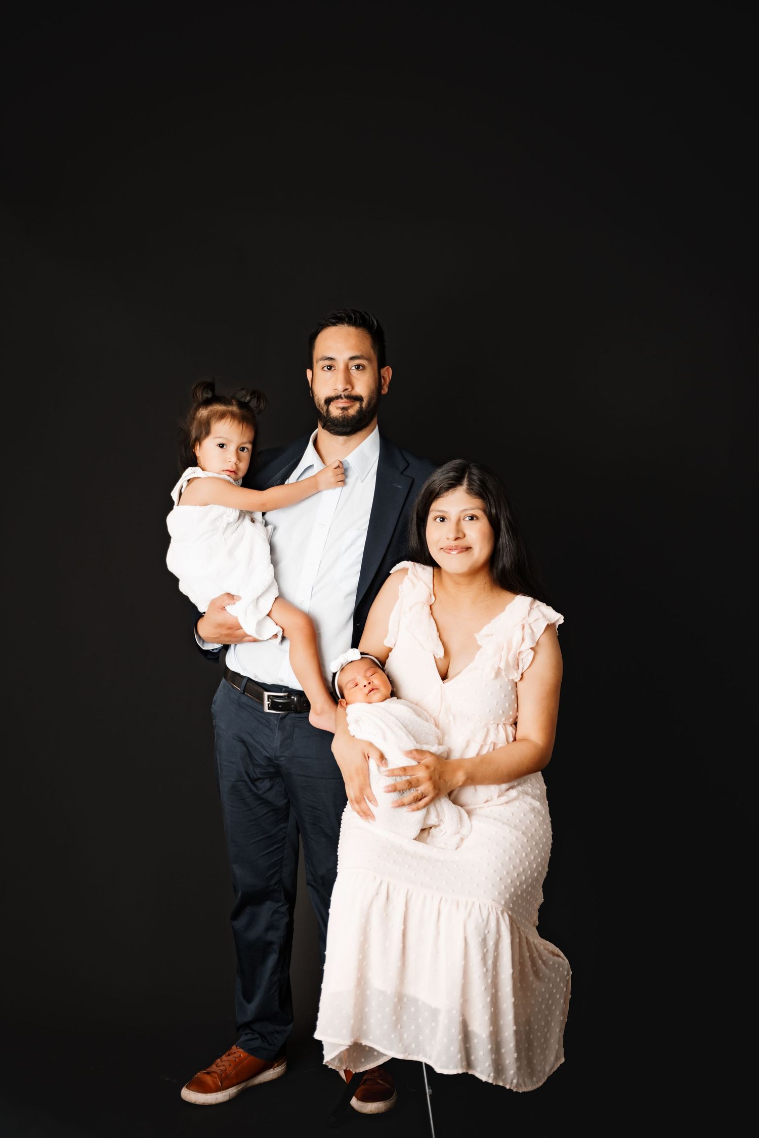 Family portrait with parents holding two young children, posed against a black studio background.