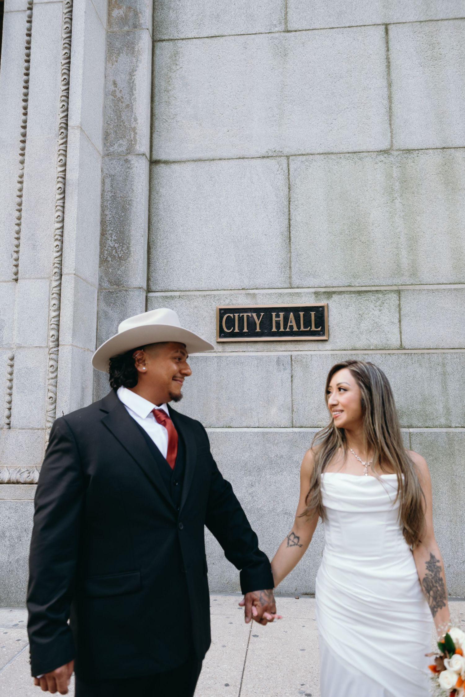 A couple in wedding attire shares a joyful moment outside City Hall.
