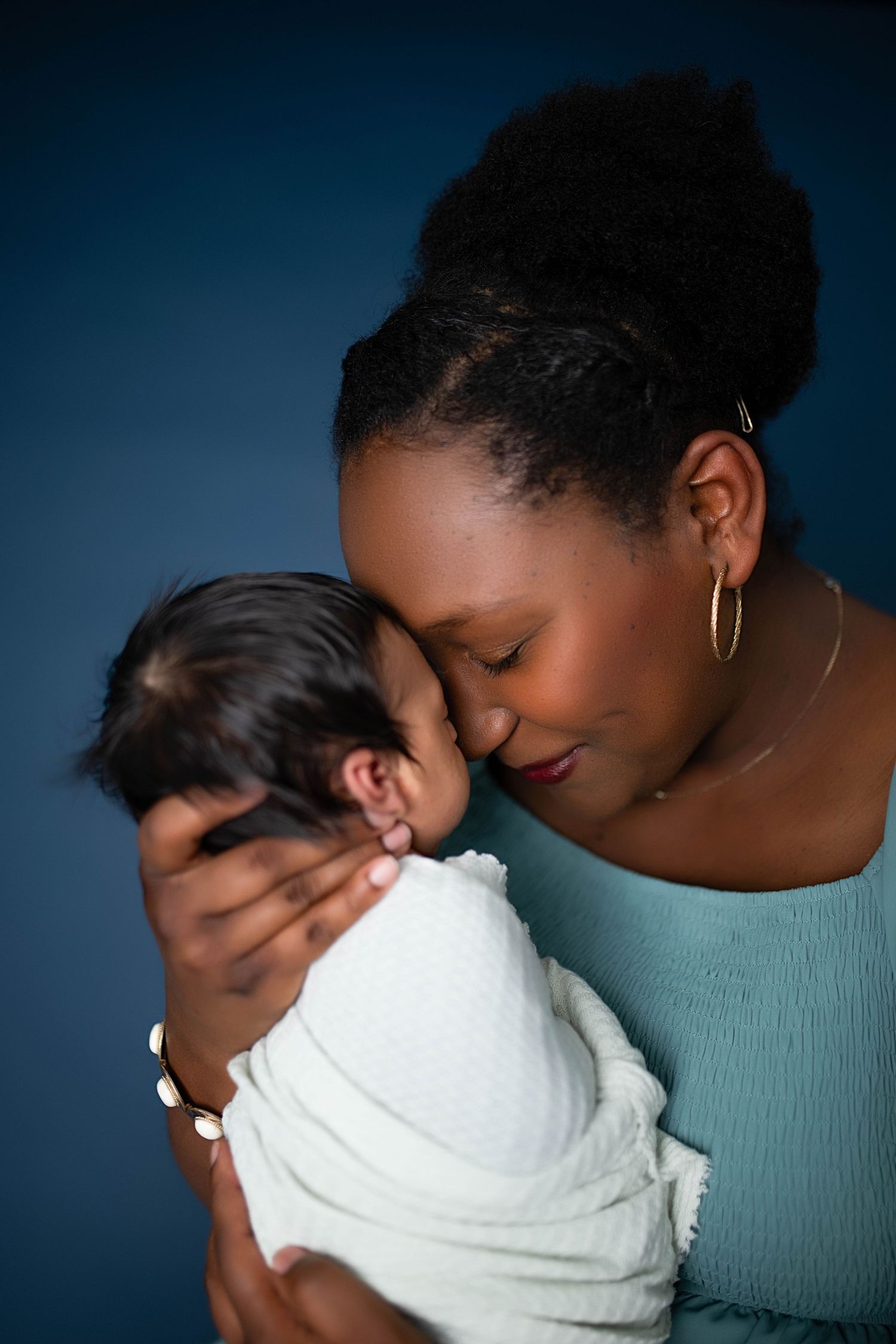 A Black mother tenderly holds her swaddled newborn baby close, touching foreheads in an intimate portrait.