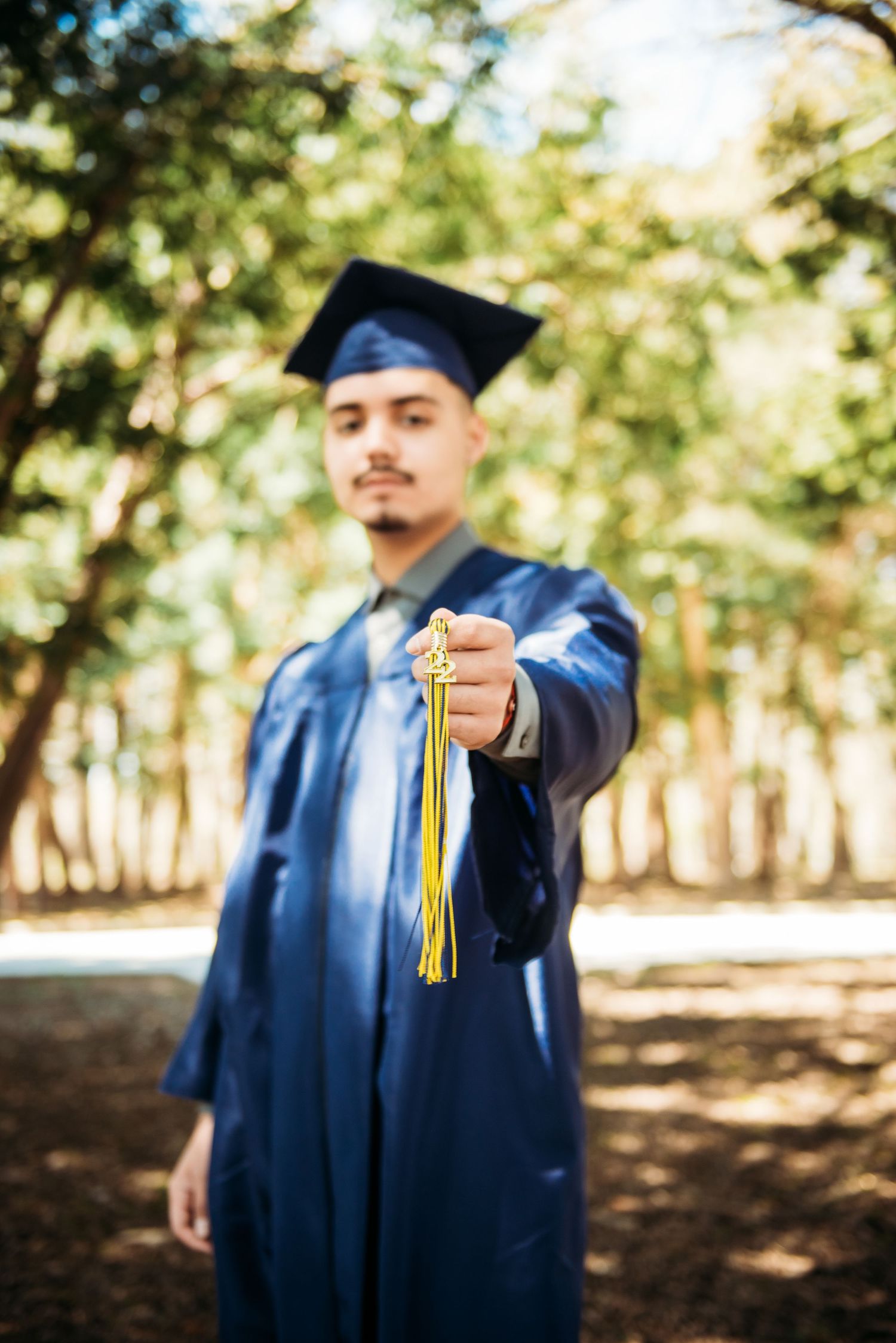 A graduate in blue cap and gown displays their tassel against a wooded background.