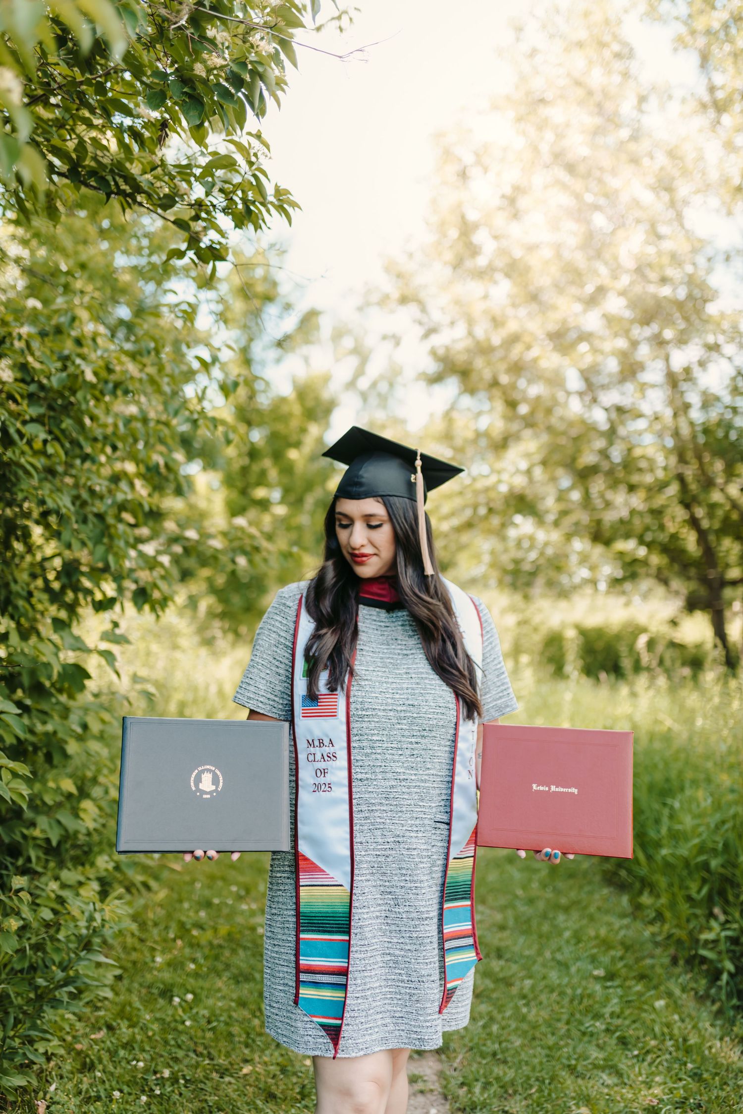 Graduate in grey dress and cap holds diploma while standing in lush green outdoor setting.