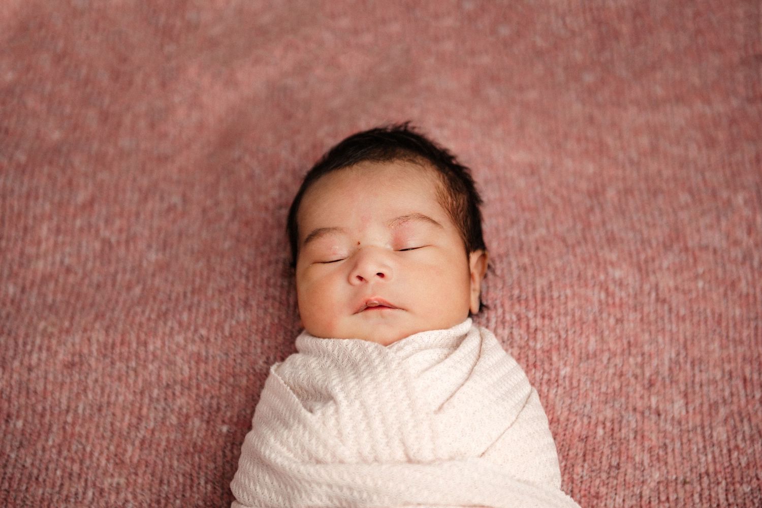 Peaceful newborn baby swaddled in white wrap sleeping on a soft pink textured background.