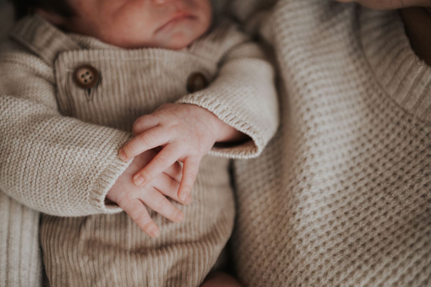A newborn baby in a beige knit romper gently holds an adult finger, capturing a tender parent-child bonding moment.
