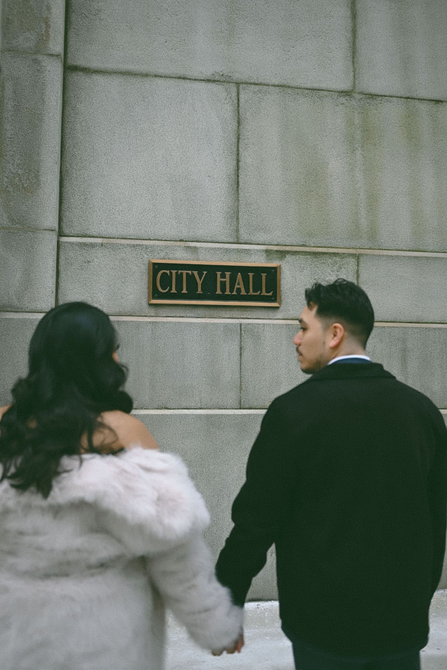 A couple playfully walks past the City Hall sign on a stone building exterior.