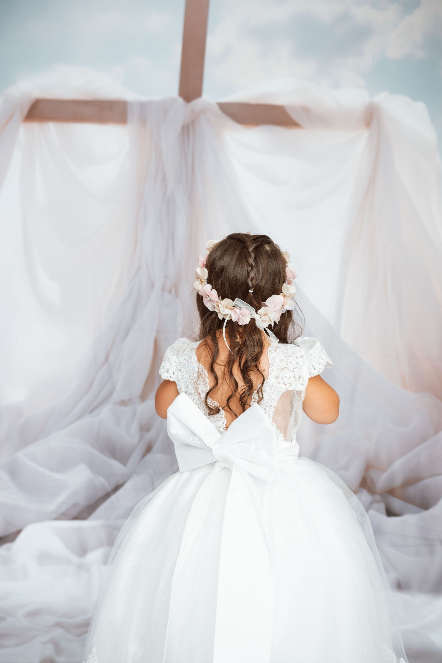 Back view of child in white dress with delicate lace details against dreamy backdrop.