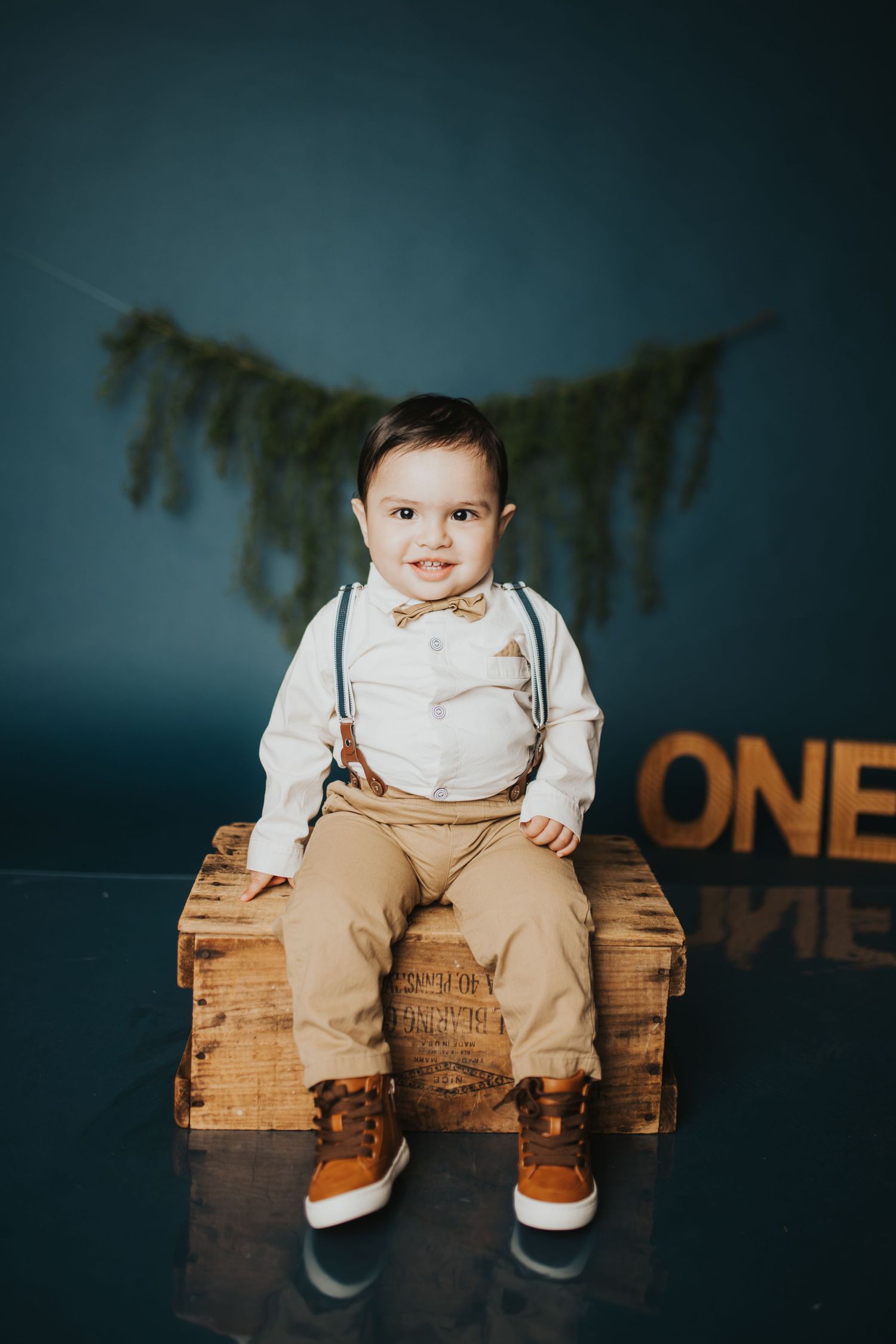 Small child in formal attire with suspenders sitting on wooden crate against dark backdrop.