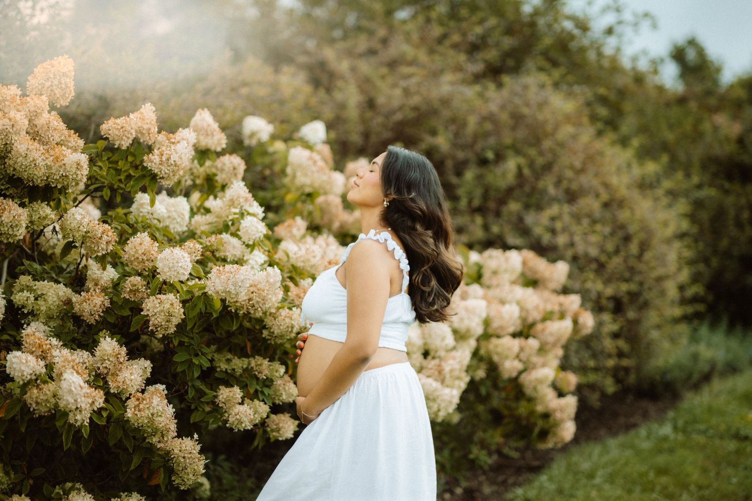 Person in white dress poses near blooming hydrangea bushes in garden setting.