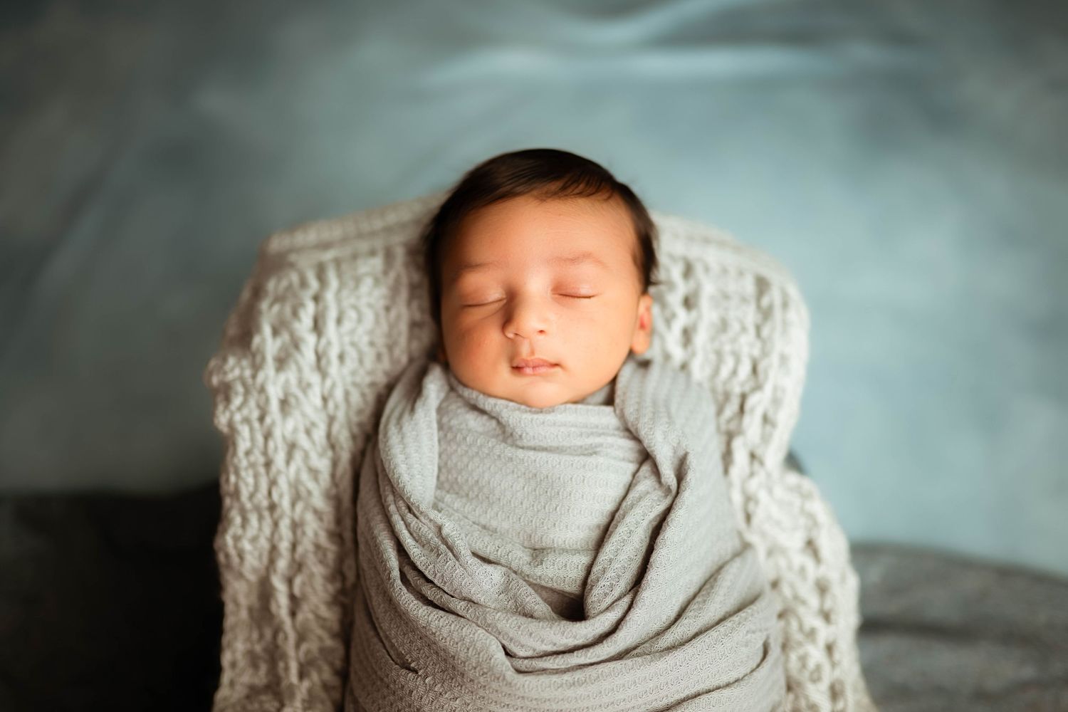 Peaceful newborn baby swaddled in gray wrap sleeping in knit basket against blue backdrop.