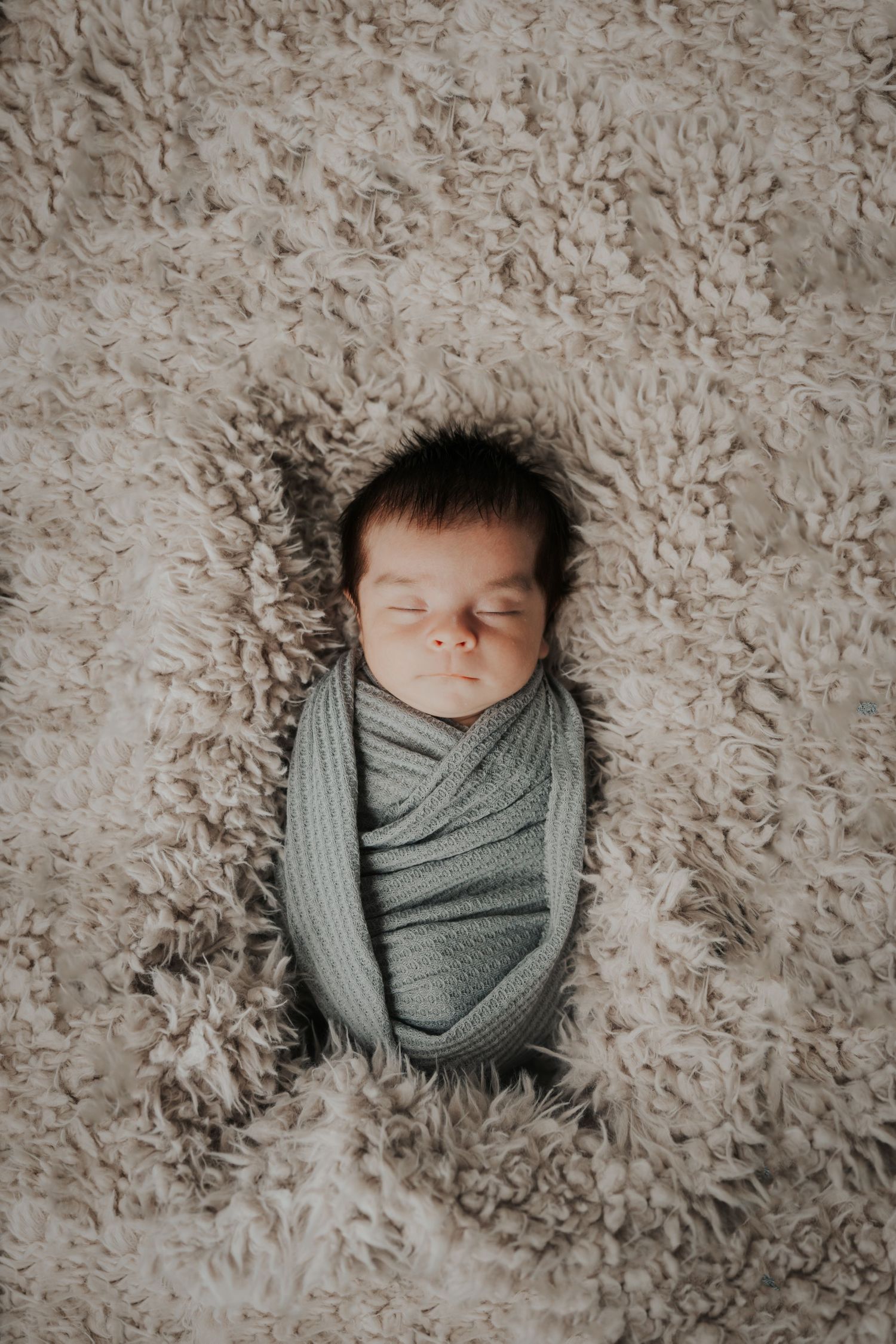 Peaceful newborn baby swaddled in a soft gray wrap, sleeping soundly on a fluffy beige textured blanket.