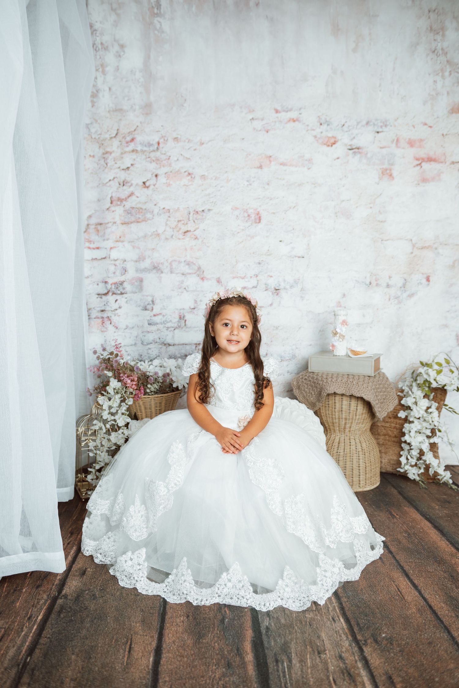 Young child in white lace dress sitting on rustic wooden floor against distressed brick wall.