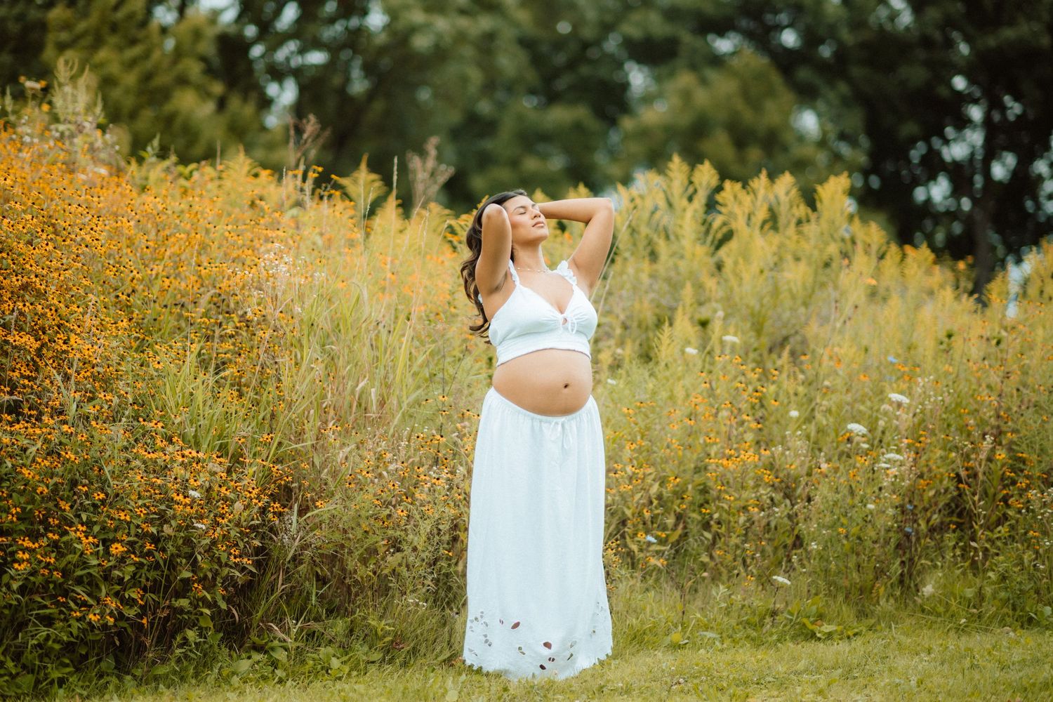 A maternity photoshoot in a field of yellow wildflowers with a white dress against a natural outdoor backdrop.