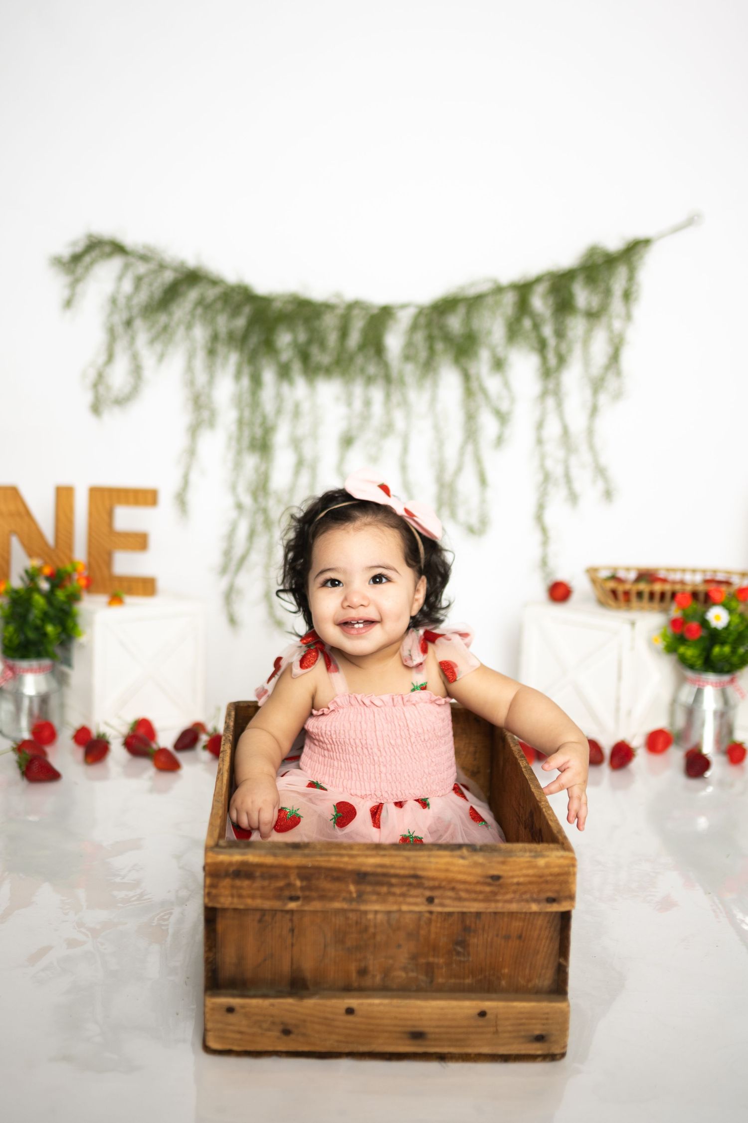 Baby sitting in rustic wooden crate during strawberry-themed photoshoot with greenery backdrop.
