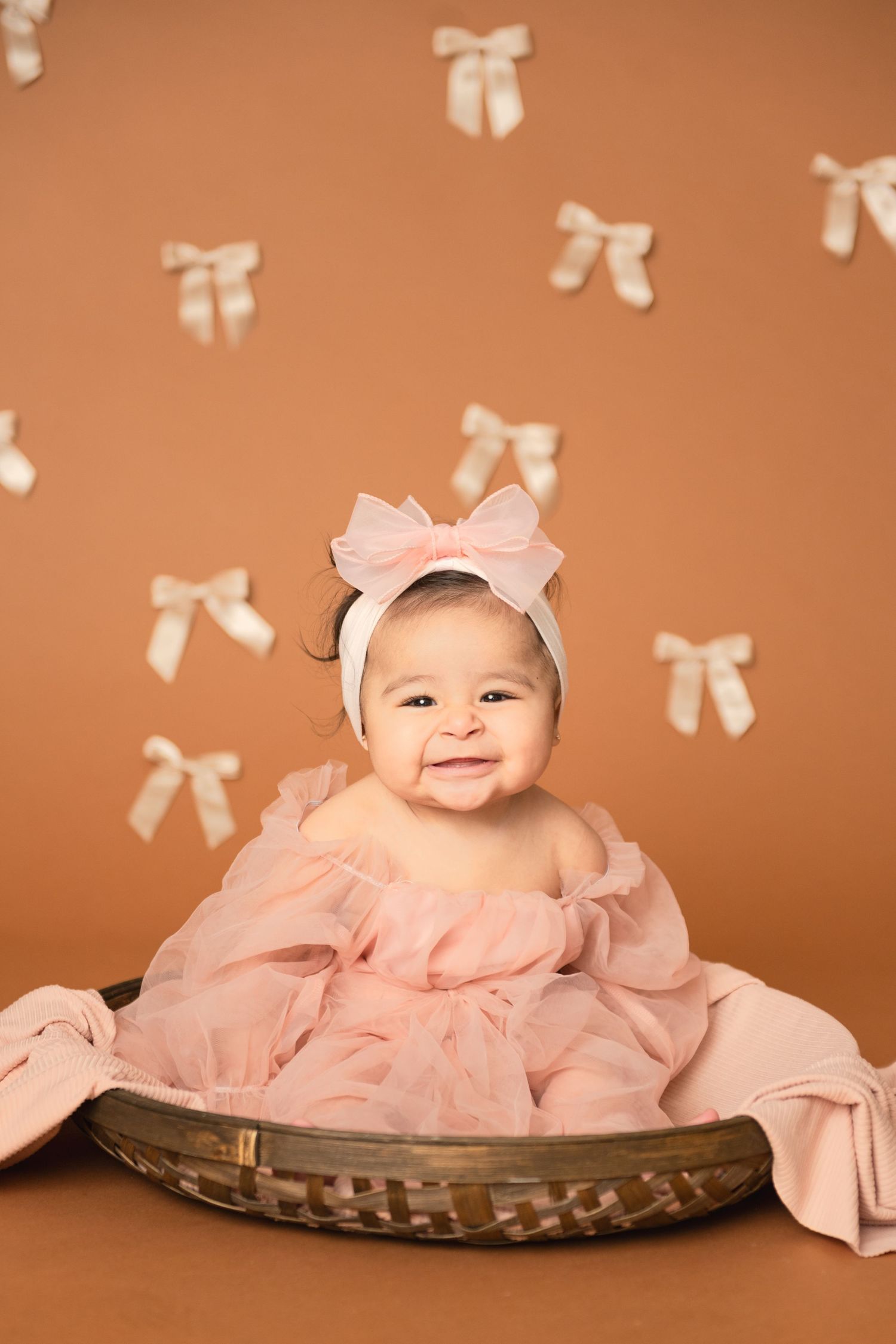 Baby in pink dress and bow sits in decorative basket against peach backdrop with bow pattern.