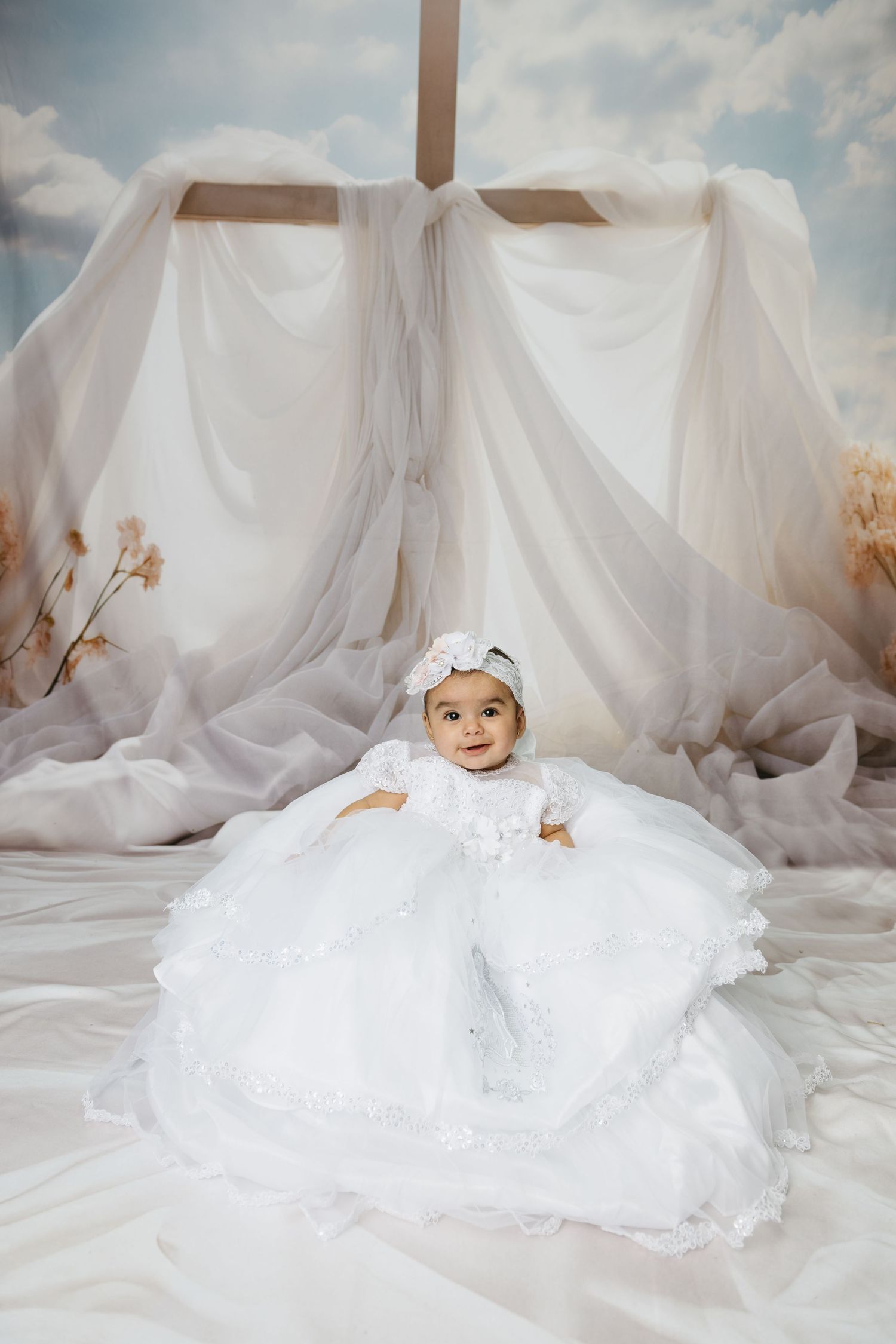 Baby wearing white christening gown lying on flowing white fabric under natural light.