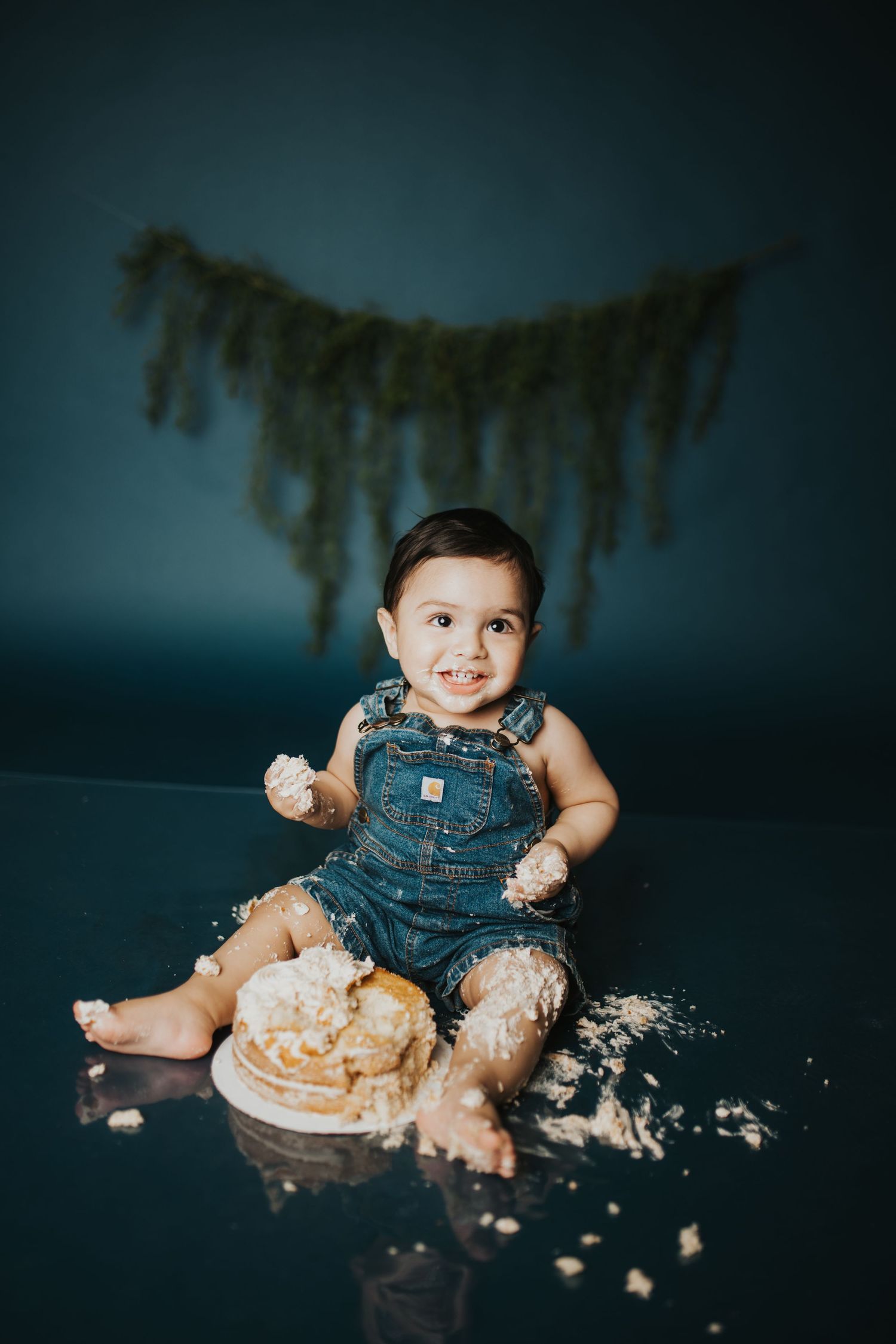 Cake smash photography with overalls and scattered frosting against dark backdrop.