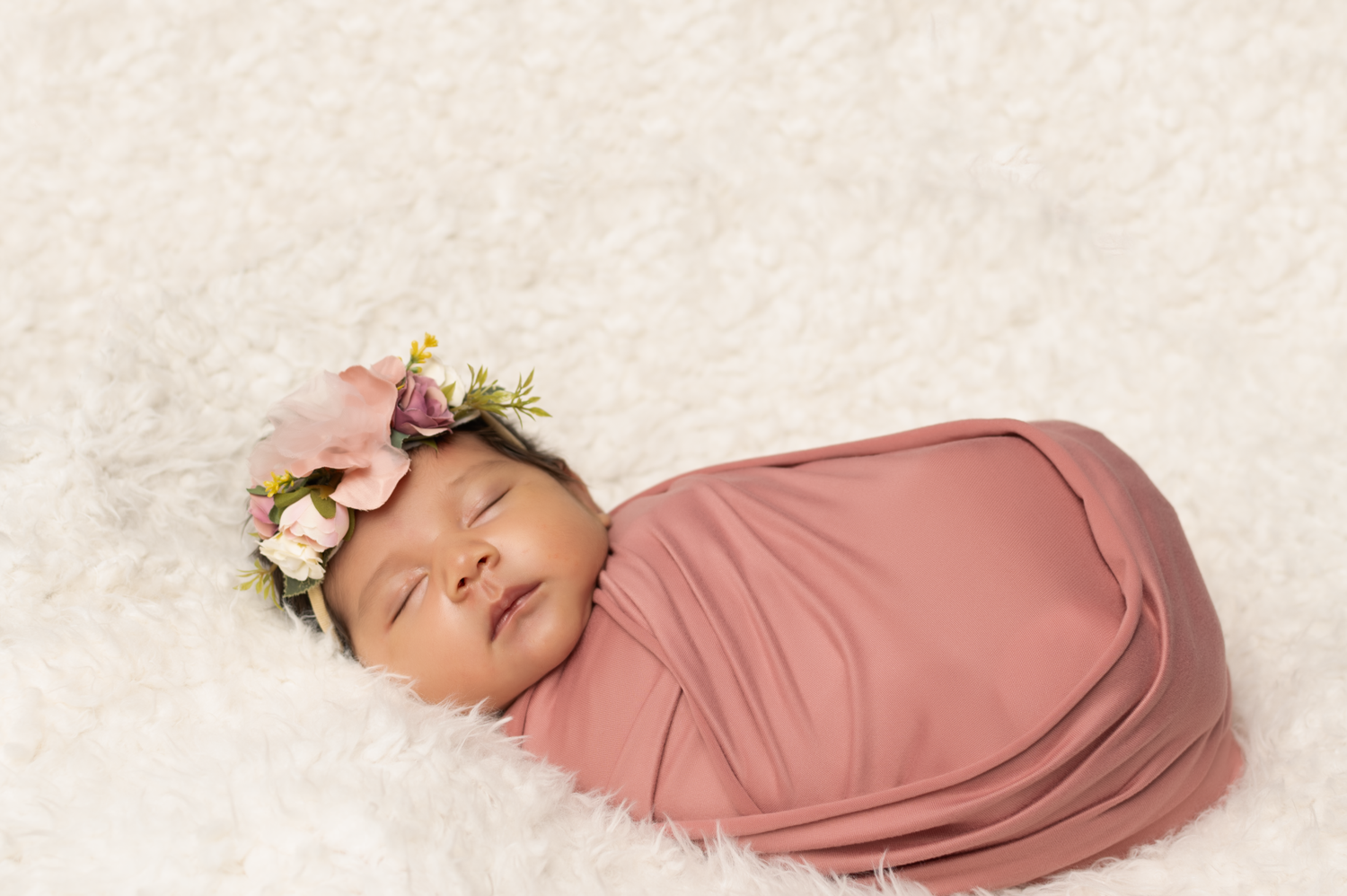 Sleeping newborn baby girl swaddled in pink wrap with floral crown on white fluffy blanket.