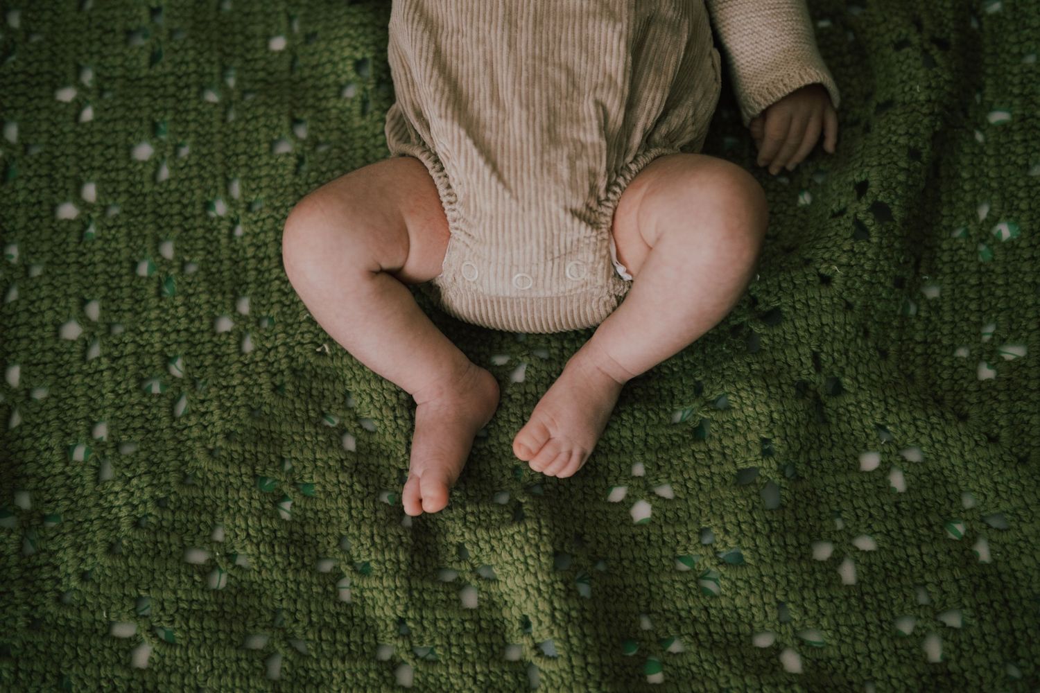Baby legs resting on a green crocheted blanket, wearing a neutral knit romper, captured from above.