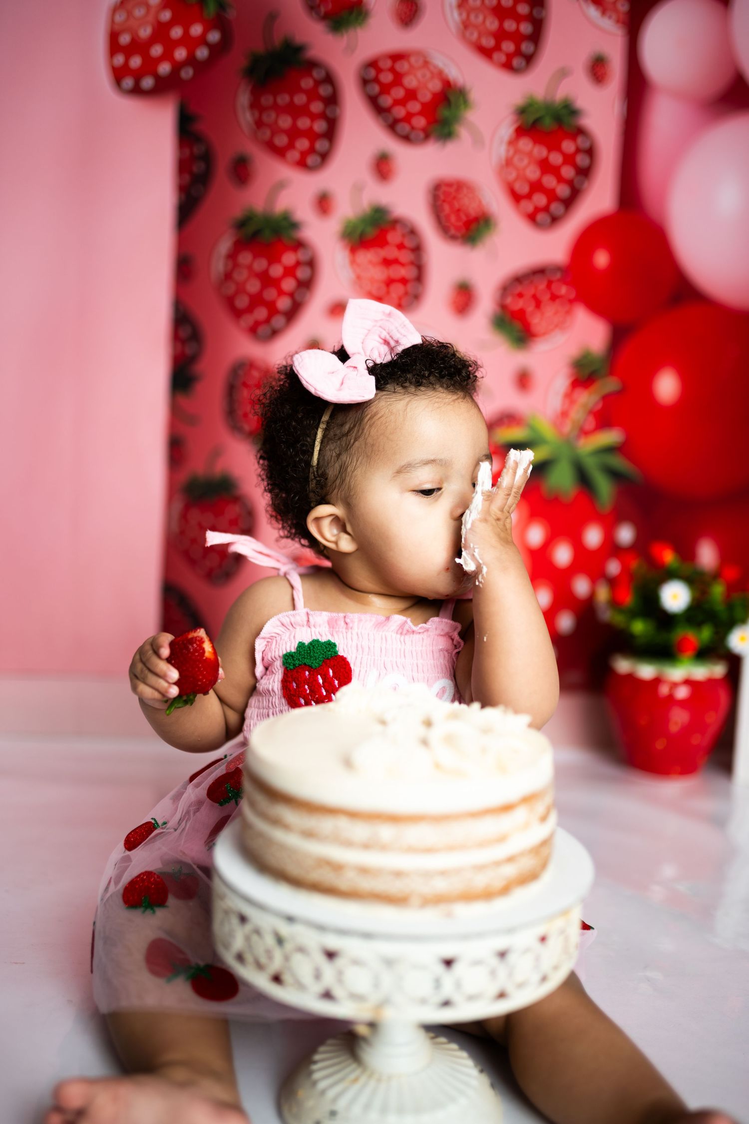 Baby reaching for cake during strawberry themed smash cake photoshoot with pink backdrop.