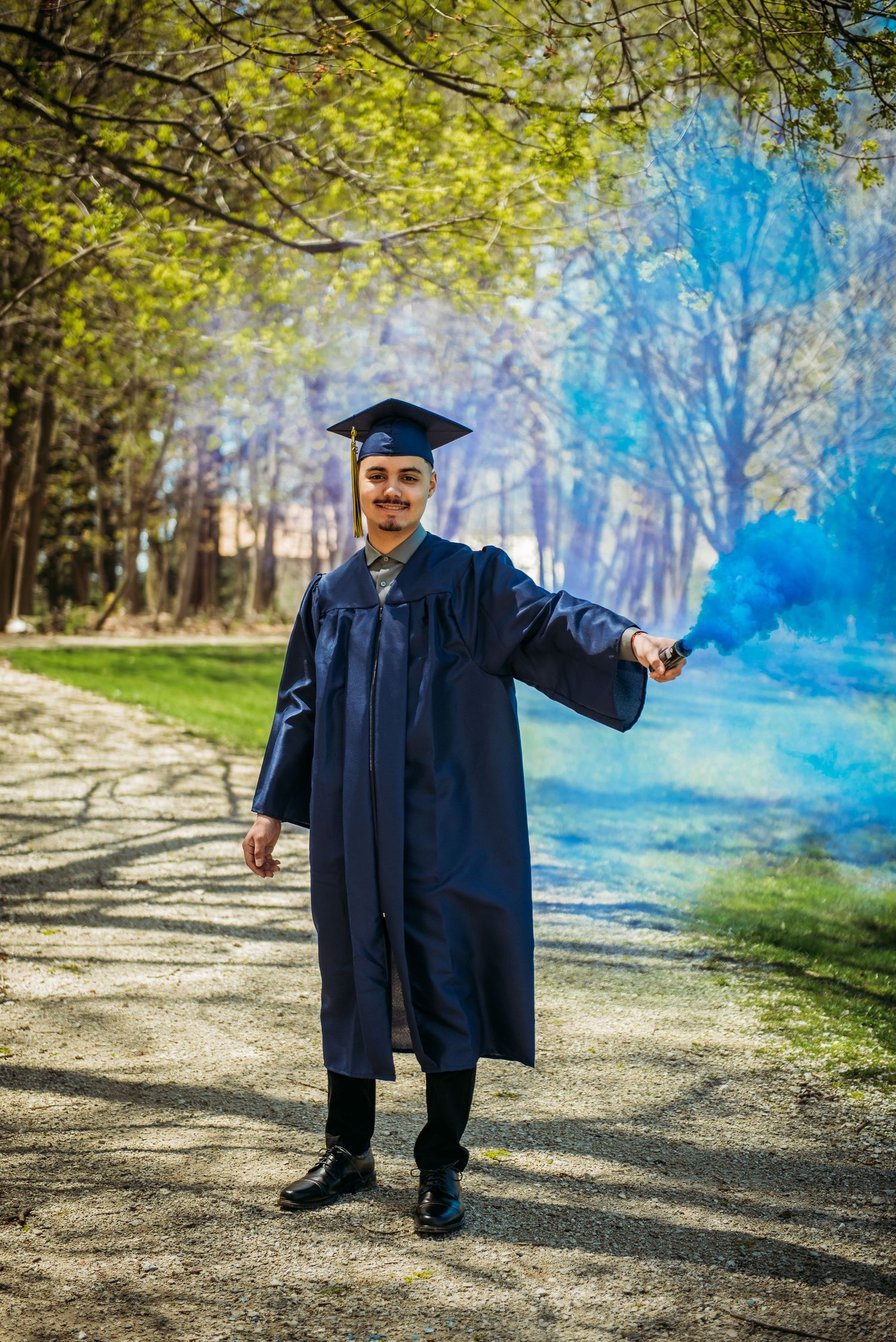 A graduate in blue academic regalia poses with blue smoke effects in a wooded setting.