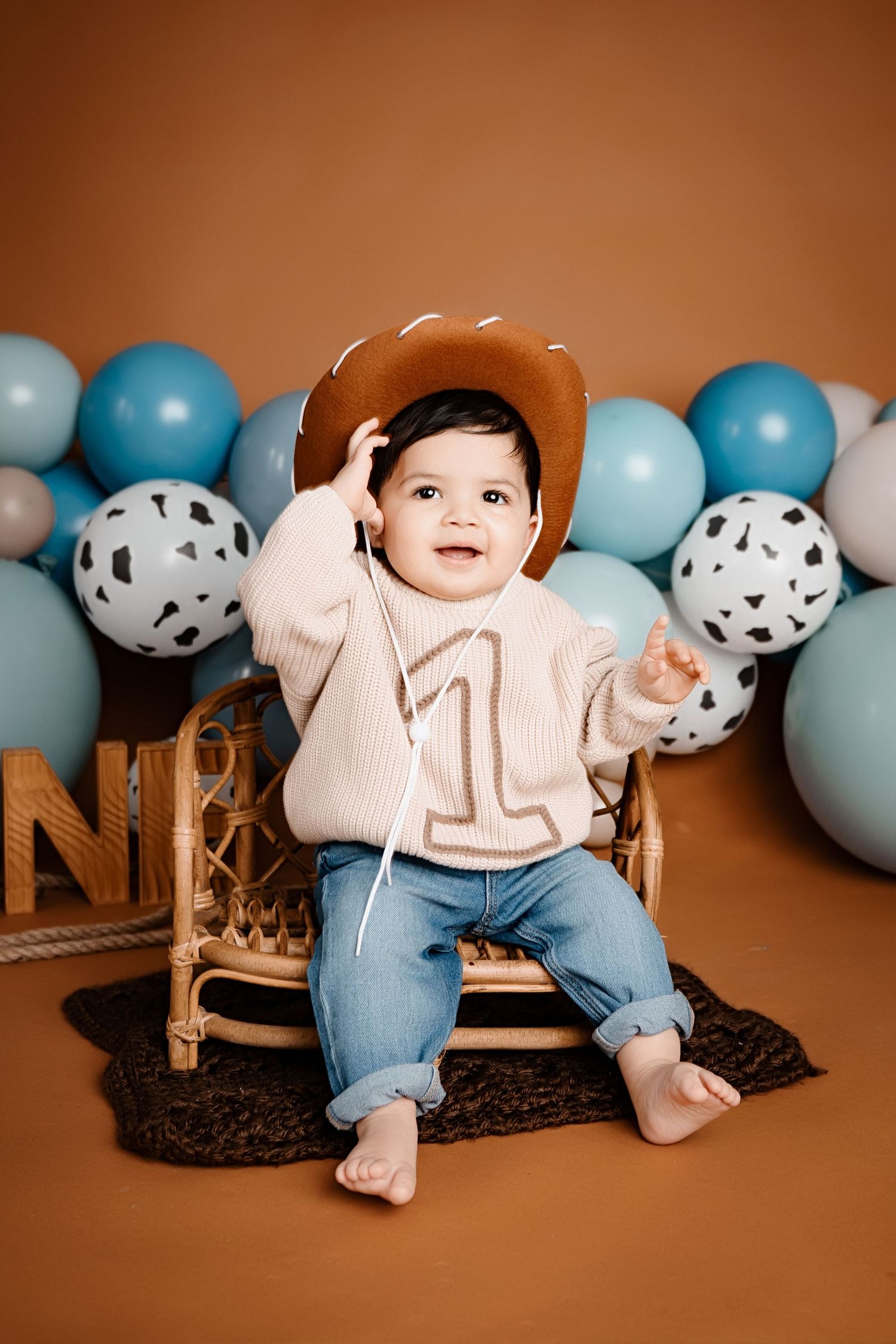 Baby in cowboy hat and sweater sits on wicker chair with blue and white balloon decorations against brown backdrop.