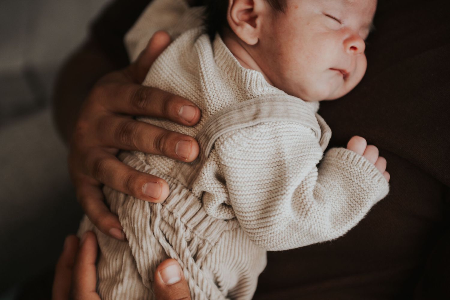 Newborn baby sleeping peacefully in cream knit outfit, cradled gently in adult hands.