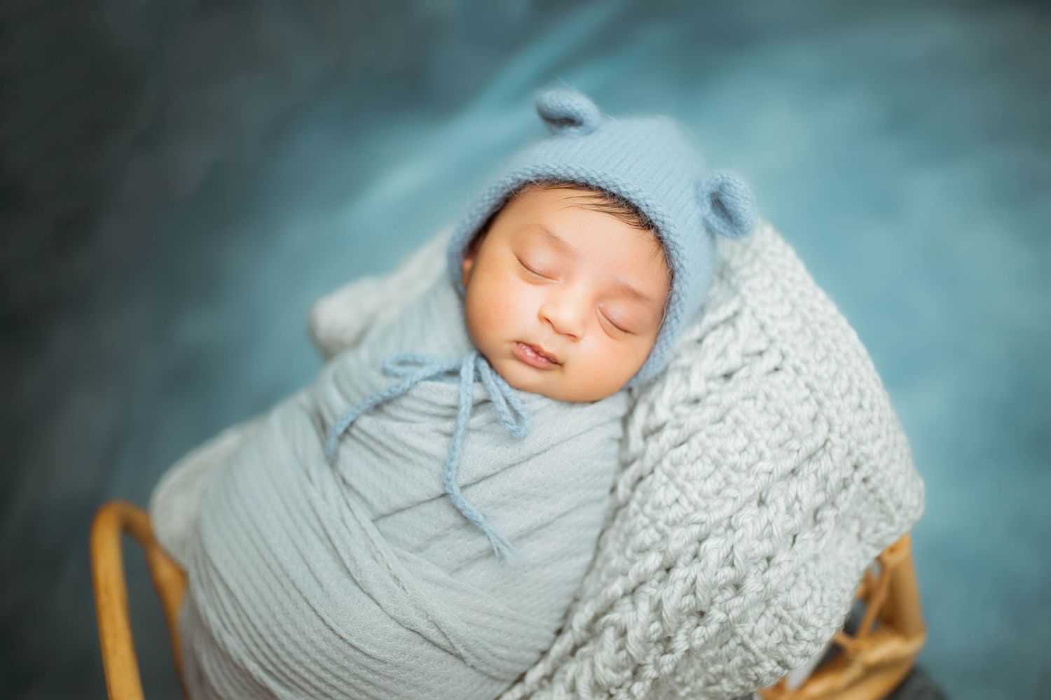 Sleeping newborn baby wrapped in blue knit blanket wearing bear hat in wicker basket on teal background.